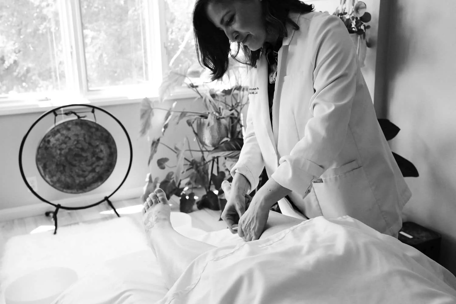 Regenerate alt text copyIconcrossIconBlue A woman in a lab coat performs painless acupuncture on a person’s leg in a bright room with plants, a gong, and women’s health focus.