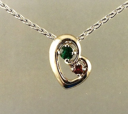 Silver necklace with a heart-shaped pendant featuring two small gemstones, one green and one red.