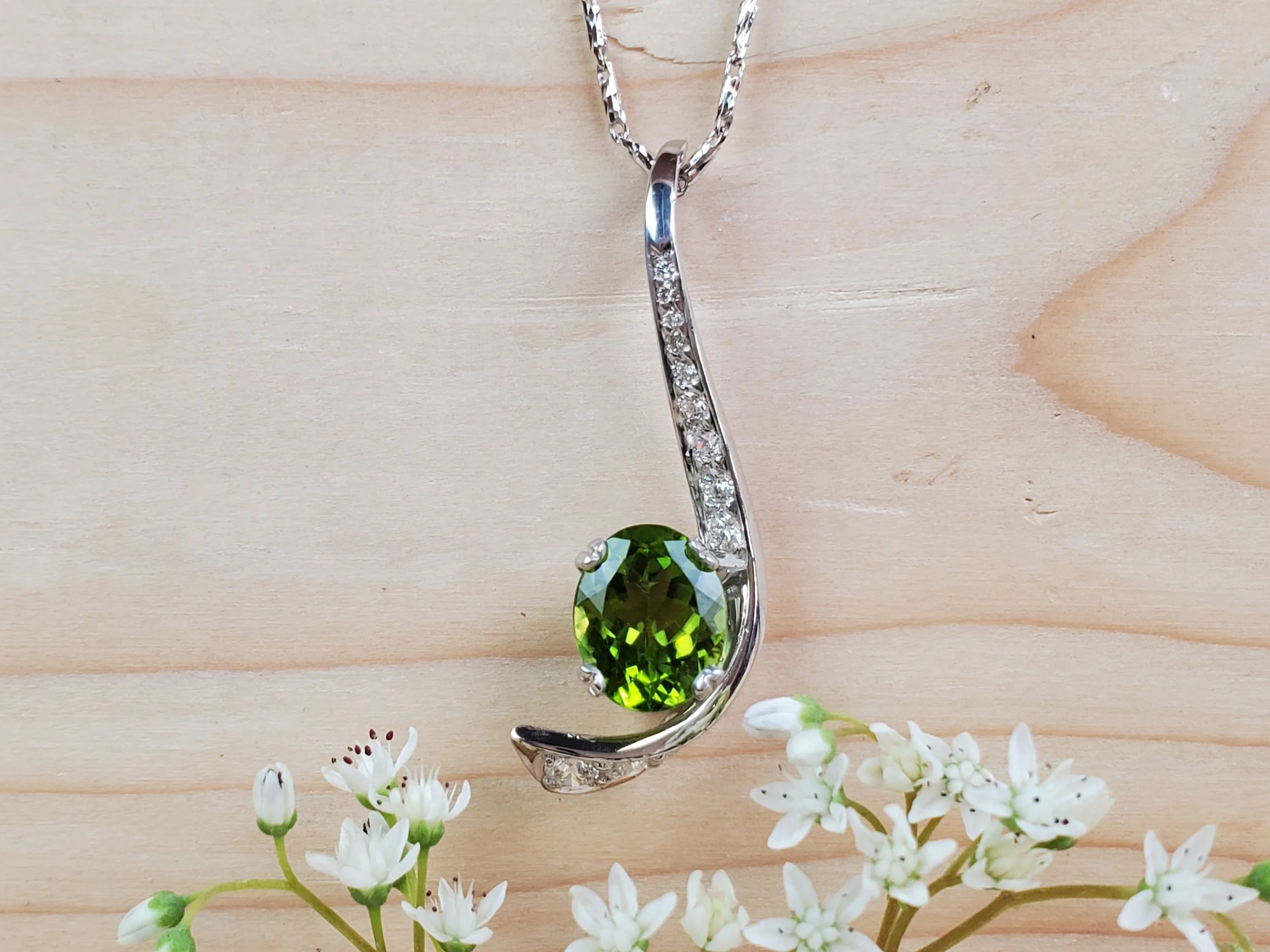 Silver necklace with a green oval gemstone pendant and small clear stones, placed on light wood surface with small white flowers at the bottom.