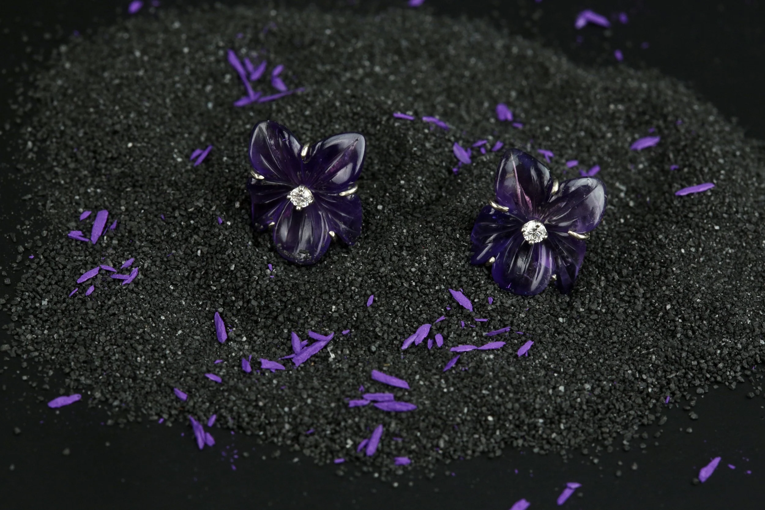 Two purple floral-shaped earrings carved out of amethyst stones with diamond centers designed and handmade by stark jewelers