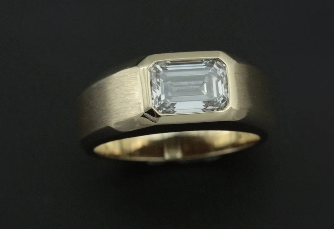 A gold ring with a rectangular emerald-cut diamond set in the center on a black background.