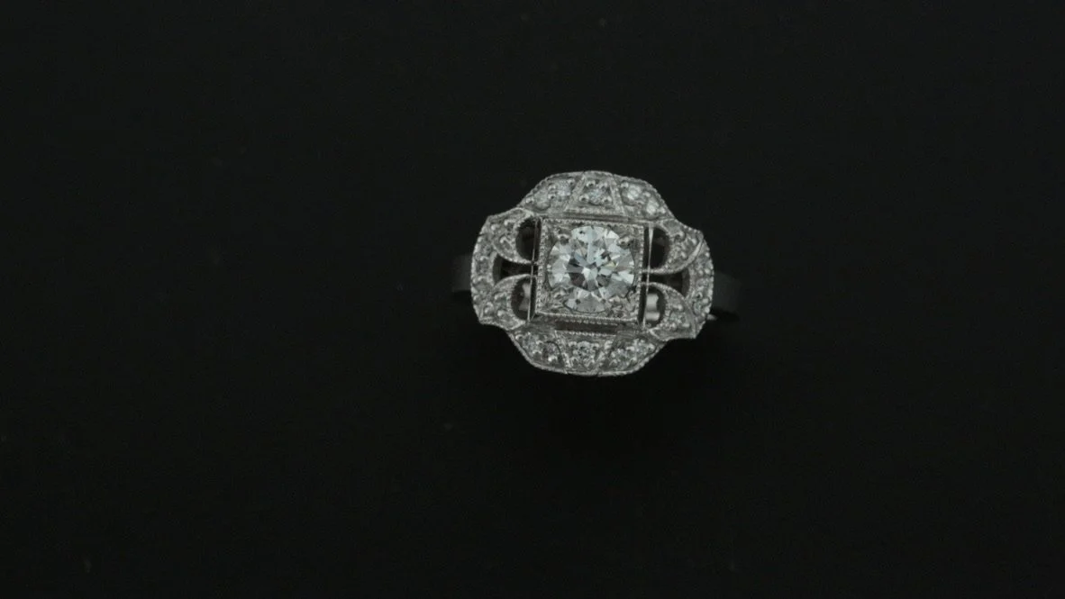 Close-up photo of an ornate diamond ring with a large center stone, set on a black background.