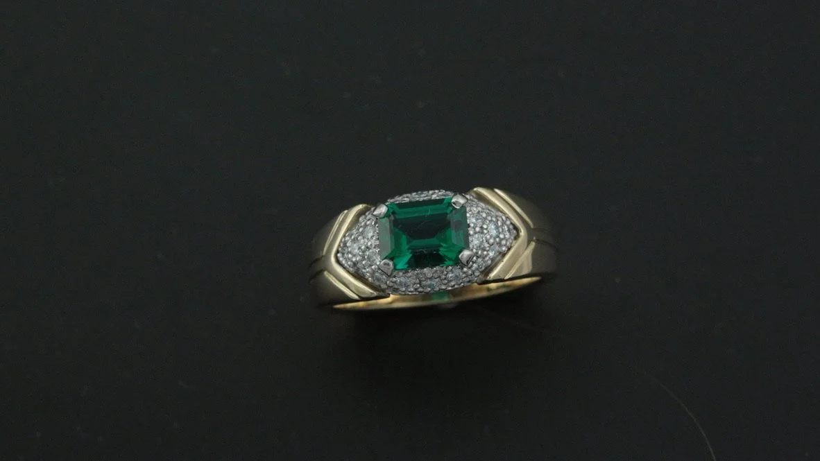 Gold ring with an emerald center stone surrounded by small diamonds on a black background.