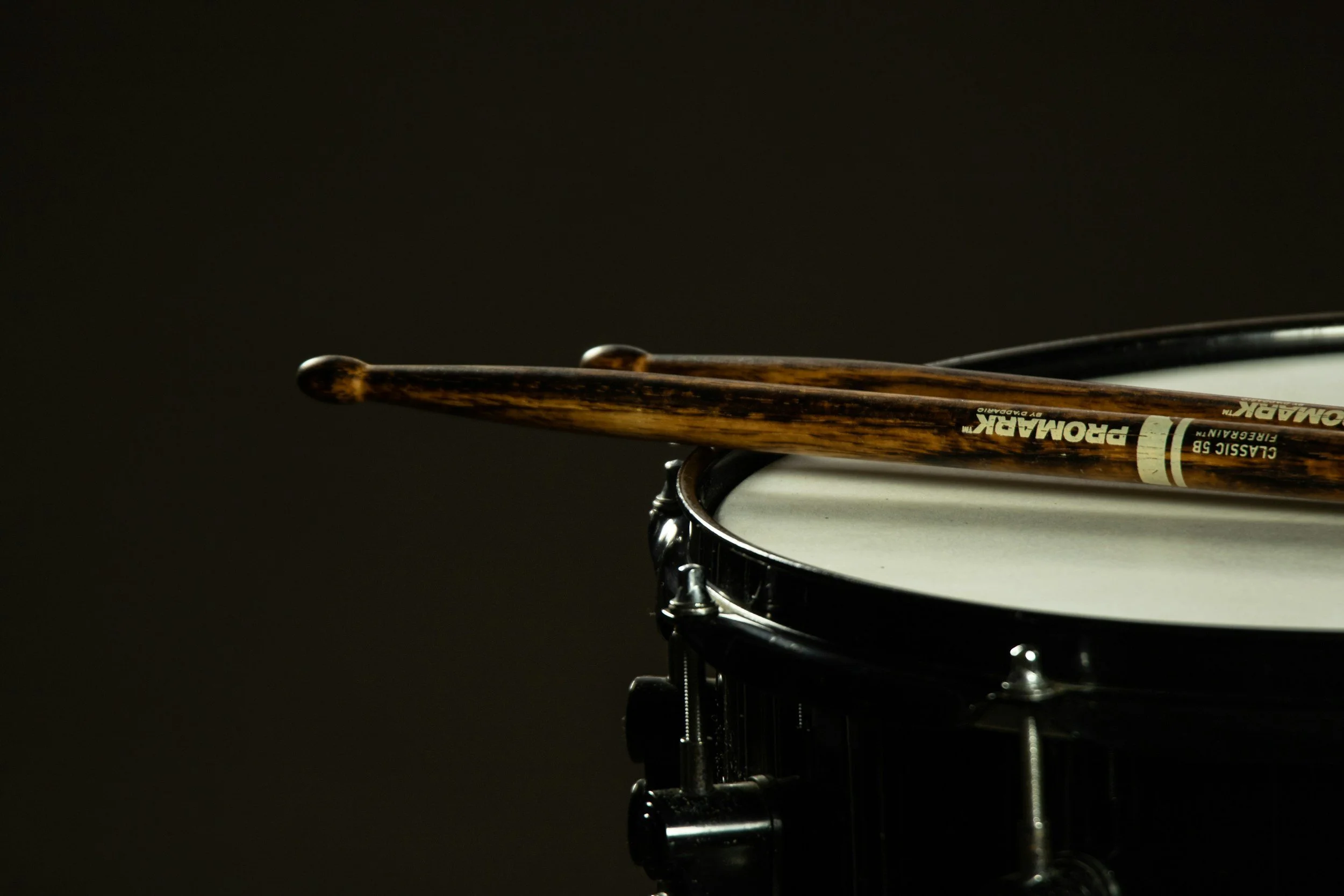 A snare drum with a pair of wooden drumsticks resting on top, set against a dark background.