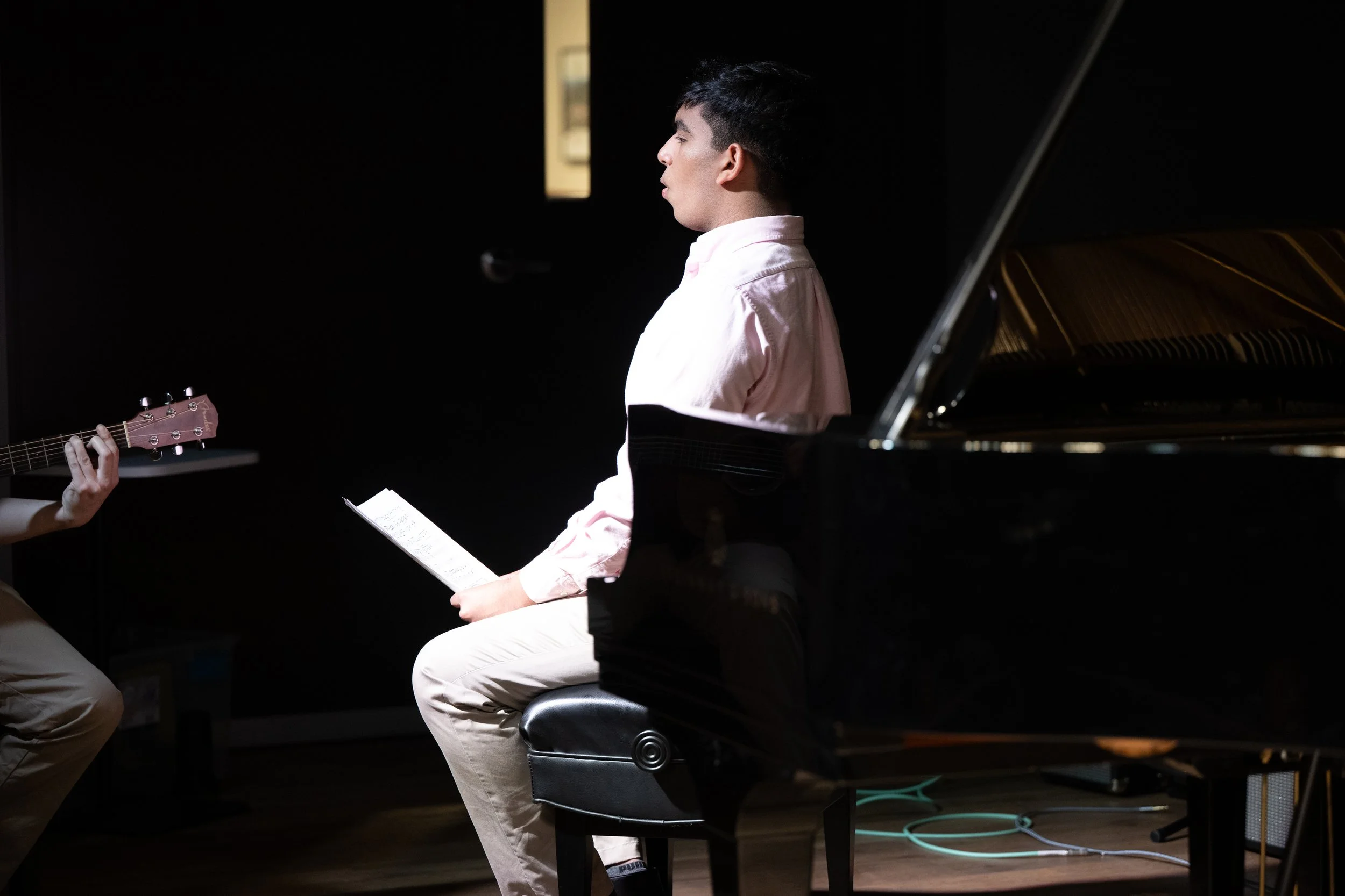 A young man in a pink shirt sitting on a black bench, holding sheet music, during a music rehearsal with a person playing guitar next to him, and a black grand piano in the foreground.