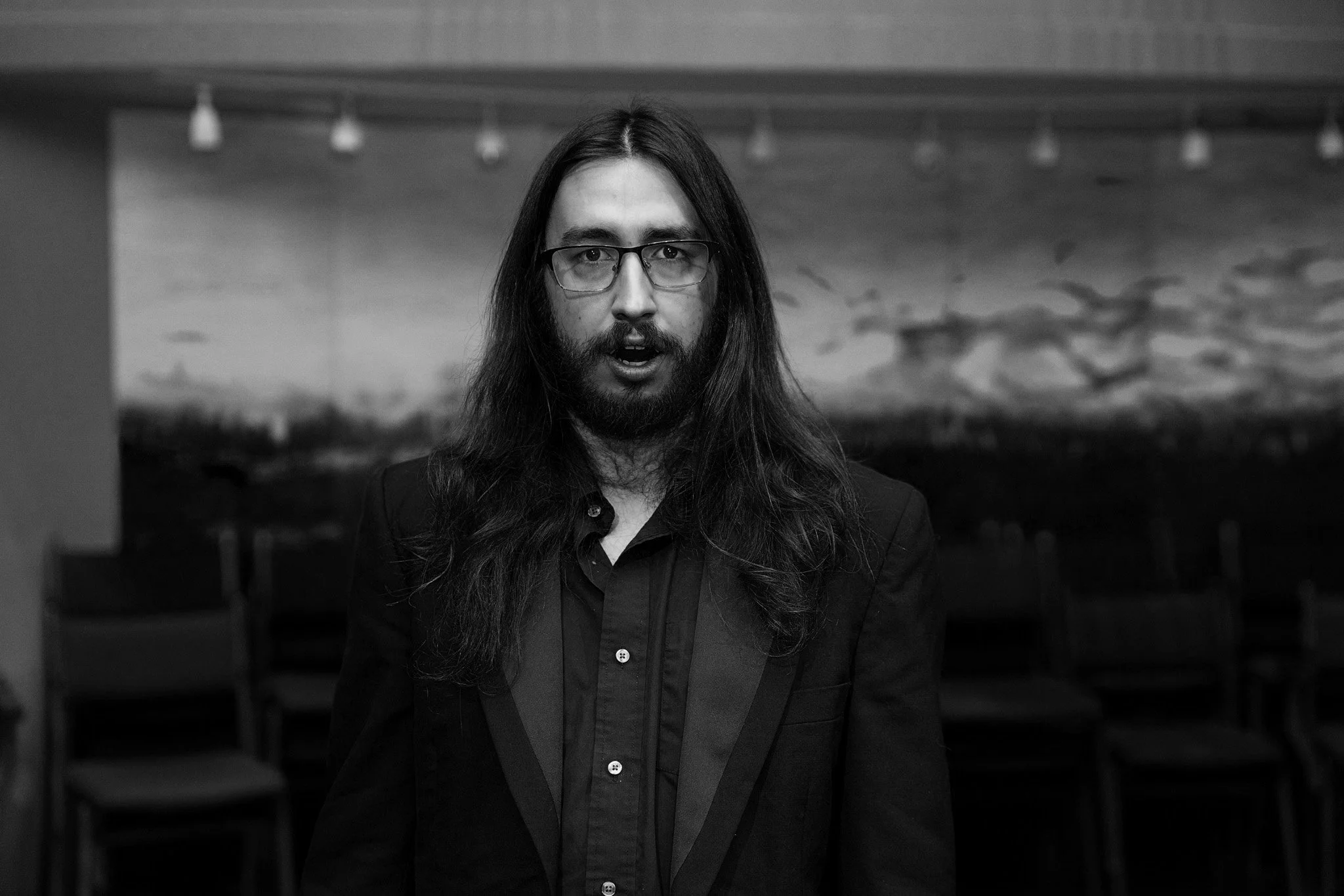 Black and white photo of a man with long hair, glasses, and a beard, wearing a dark blazer and shirt, standing in front of chairs and a mural or photograph of a landscape with clouds.