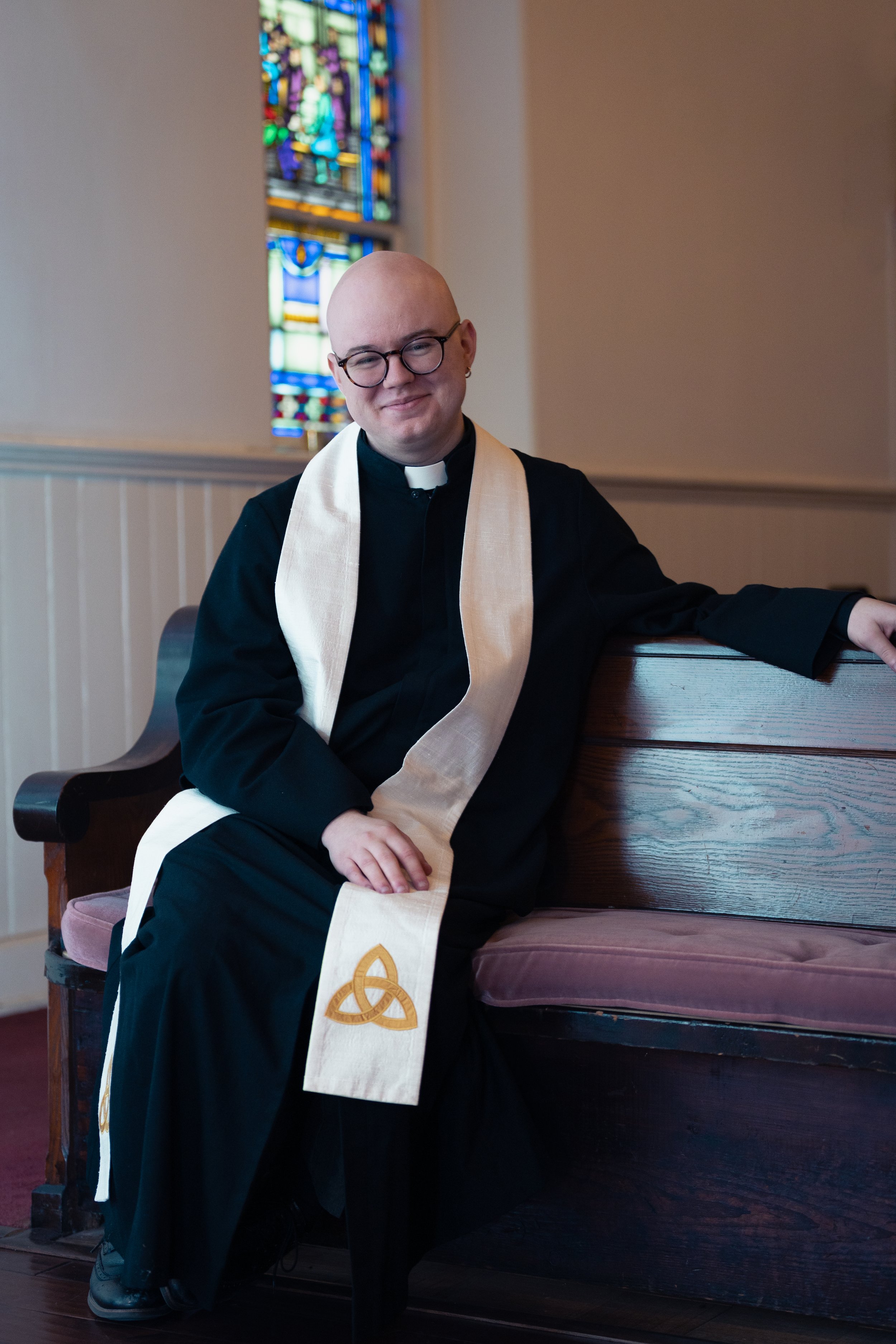 Michael Cuppett sitting on a church pew inside a church, with a stained glass window in the background, wearing black robes, a white clerical collar, glasses, and a white stole with a gold knot design.