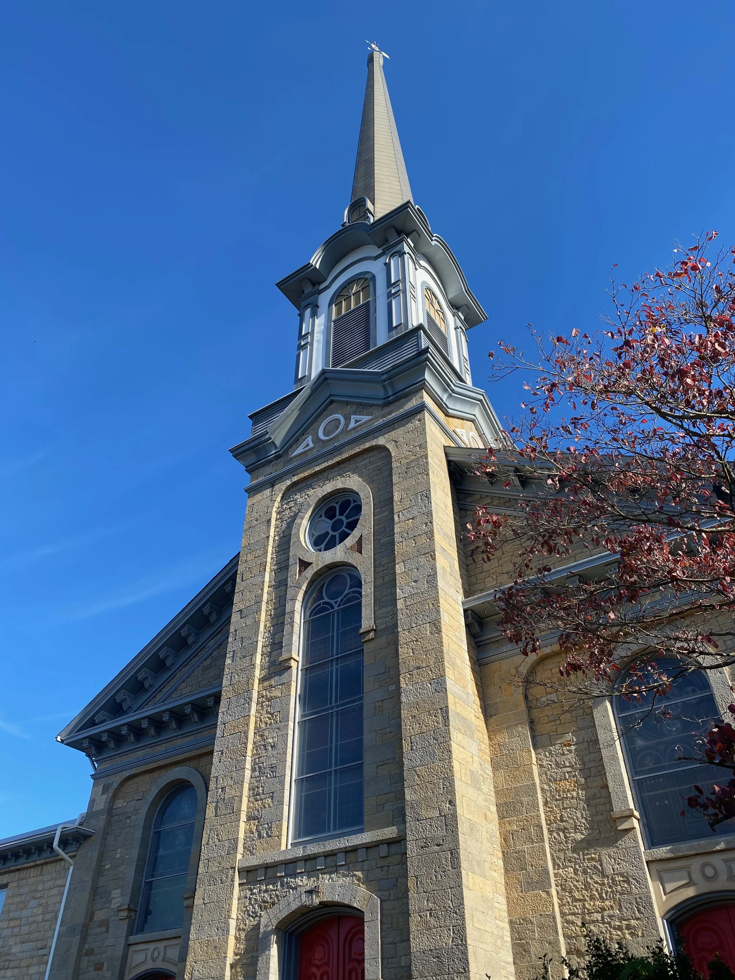 Low-angle view of a stone church with tall steeple, arched windows, and a tree with red leaves, under a clear blue sky.