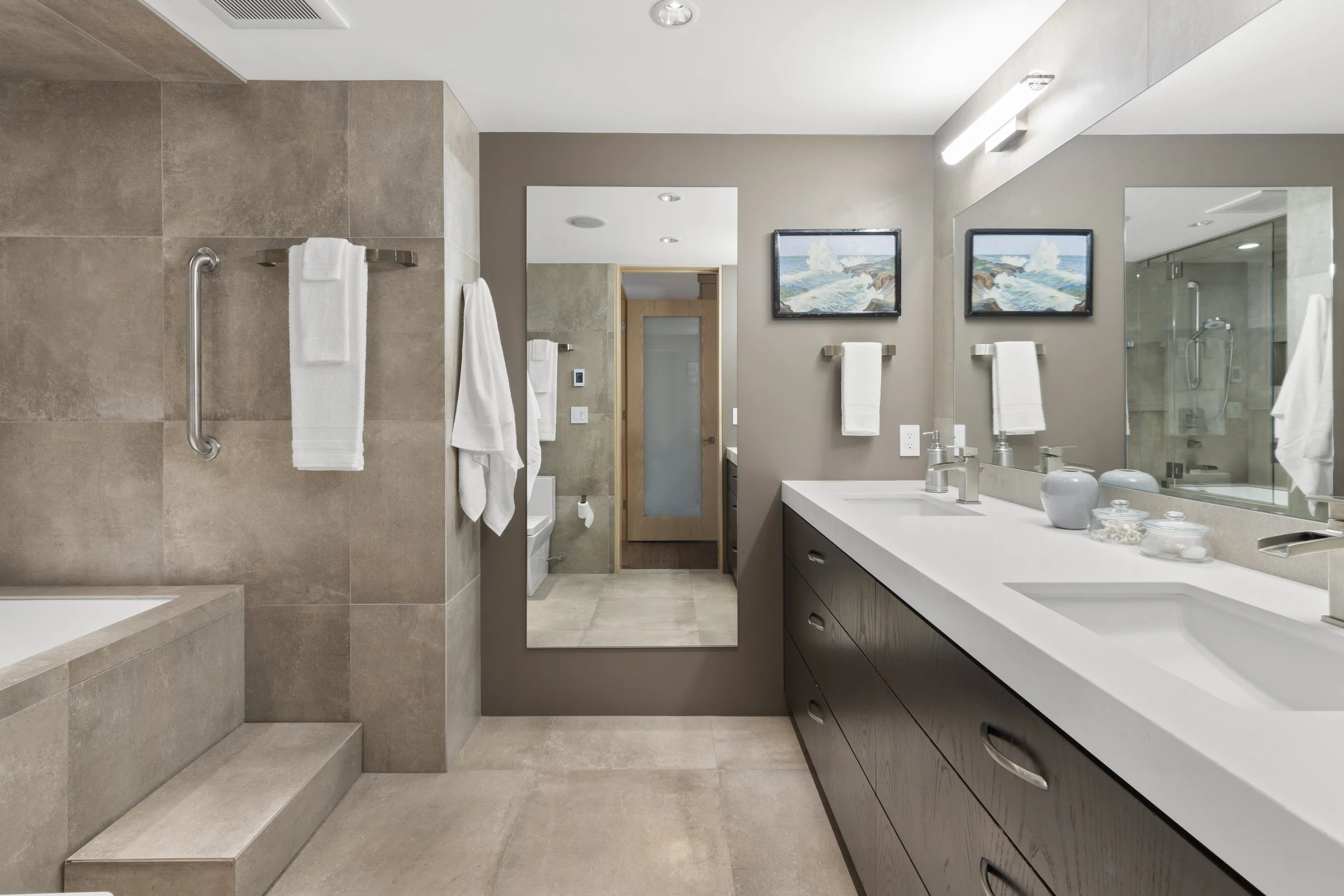 Modern bathroom with double sink vanity, large mirror, and gray tile accents. Features a bathtub with overhead towel rack and white towels. Neutral color palette with decorative wall art and sleek fixtures.
