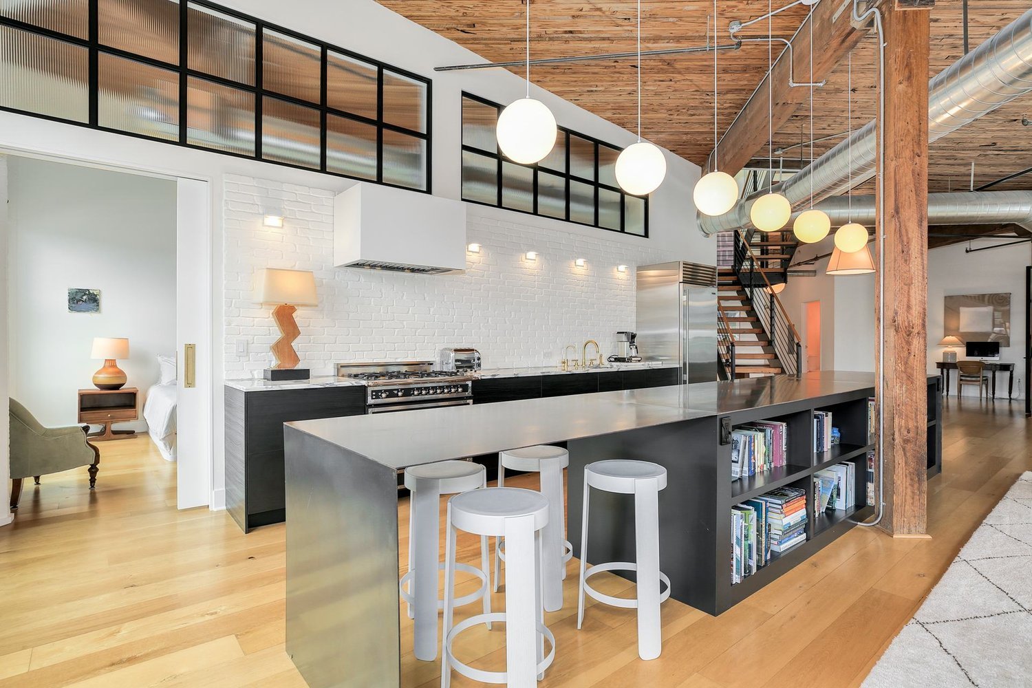 Modern kitchen and living area with wooden floors, exposed beams, a large island with bar stools, pendant lights, and a staircase. The kitchen has a stainless steel range, white brick backsplash, and open shelving. A sliding door leads to a bedroom with a lamp and artwork.