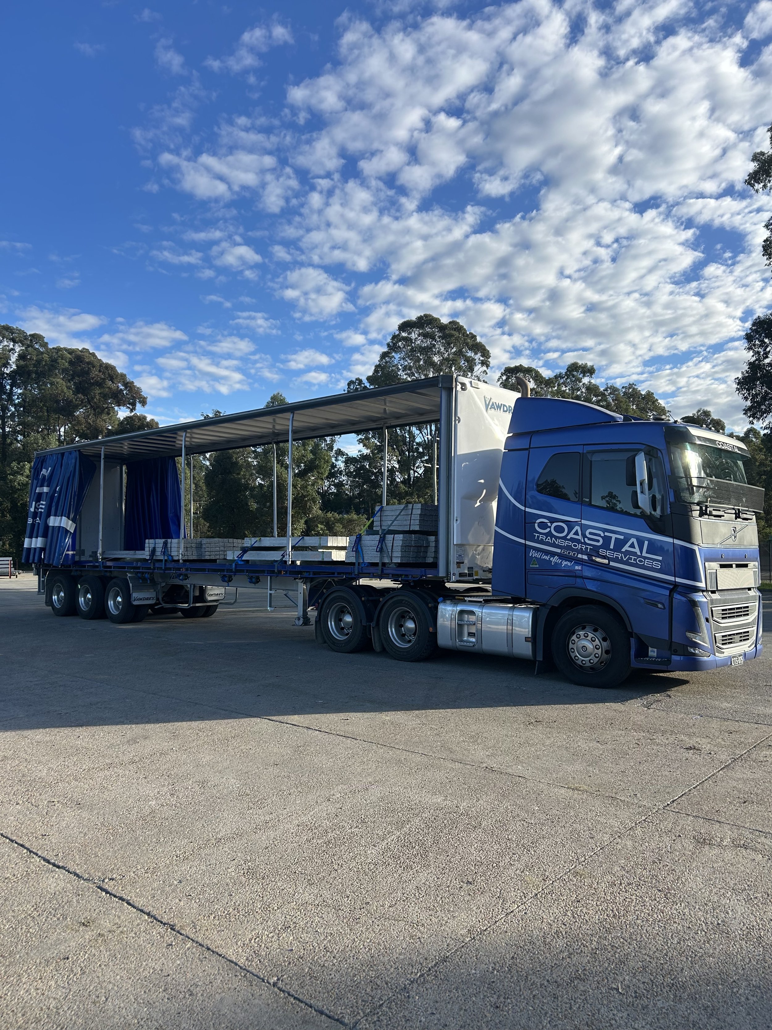 Blue semi-truck with a covered trailer parked on a paved lot under a partly cloudy sky, with trees in the background.