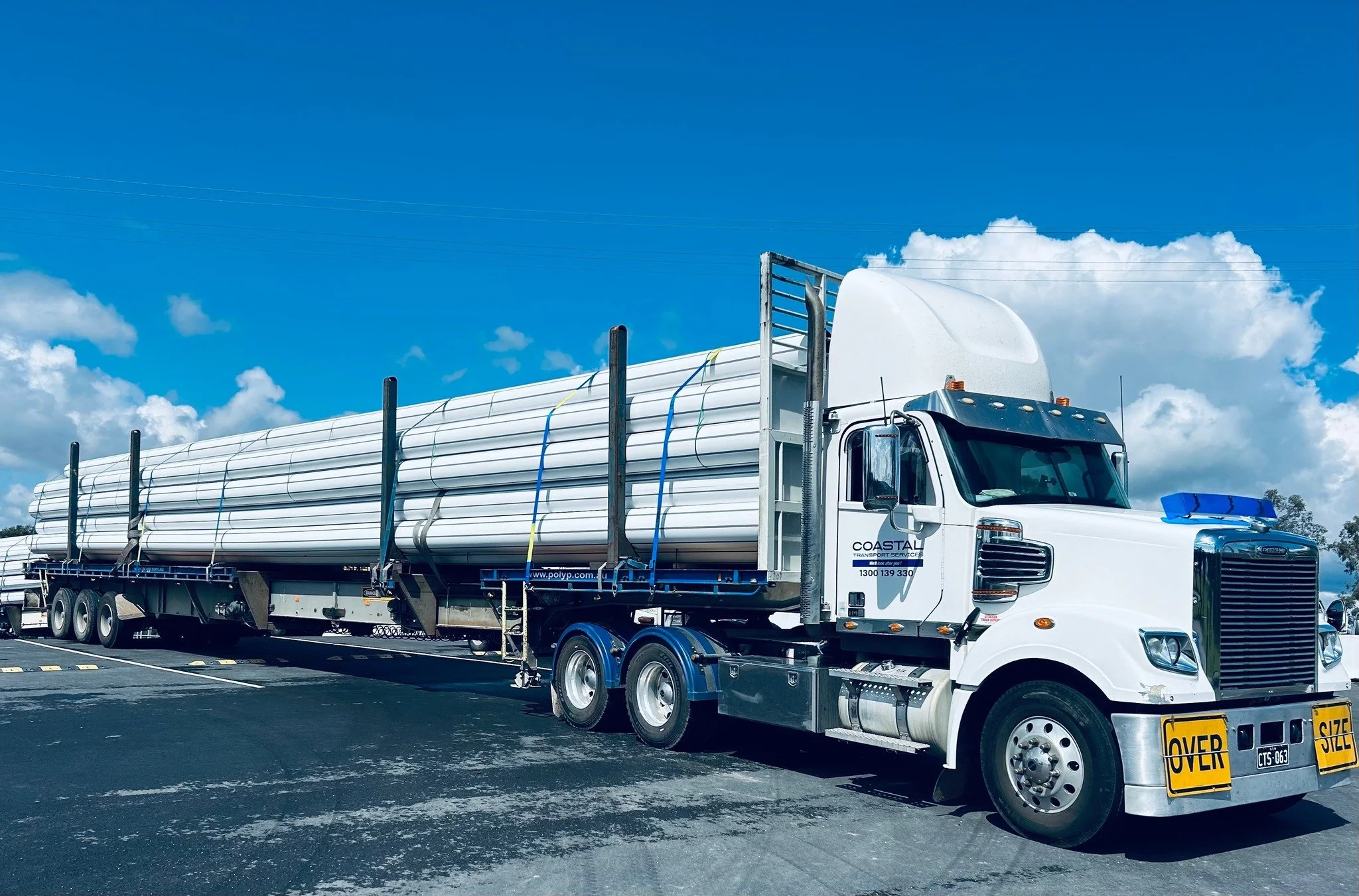 White semi-truck transporting large white pipes on a flatbed, parked on a paved lot under a partly cloudy blue sky.