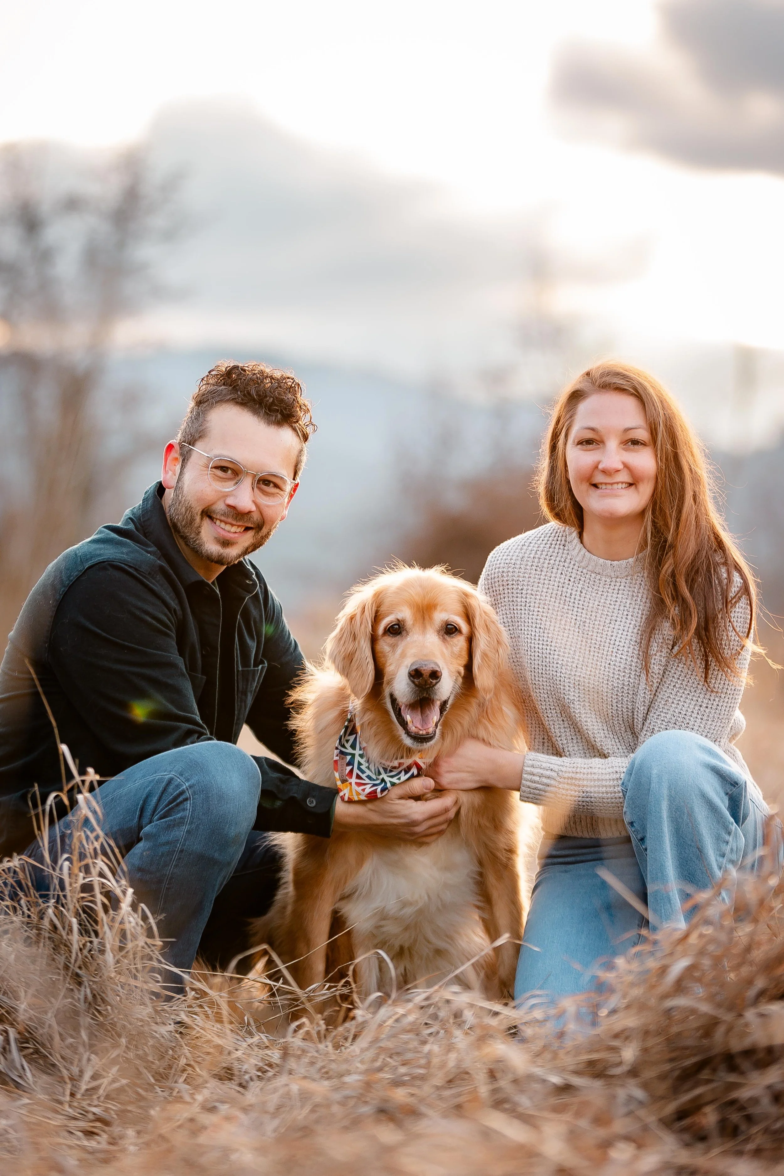 A smiling man and woman kneeling outdoors with a golden retriever dog, surrounded by tall dry grass, during sunset.