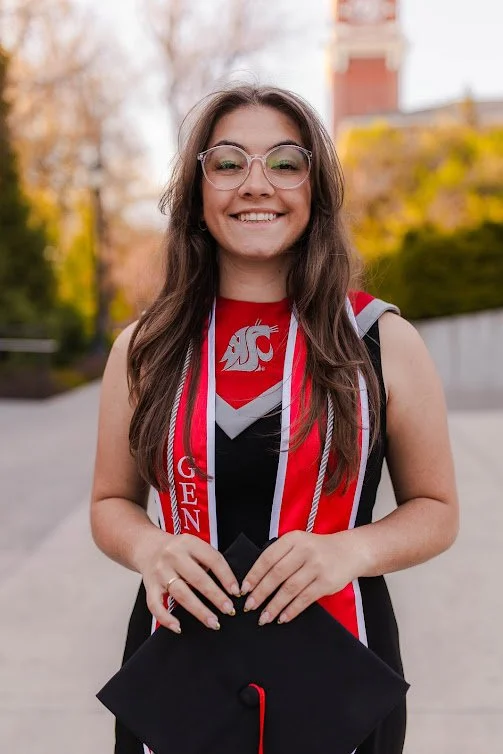 A young woman smiling outdoors during graduation day, wearing glasses, a gown, and a red and black dress with a Washington State University logo, holding a graduation cap in her hands.