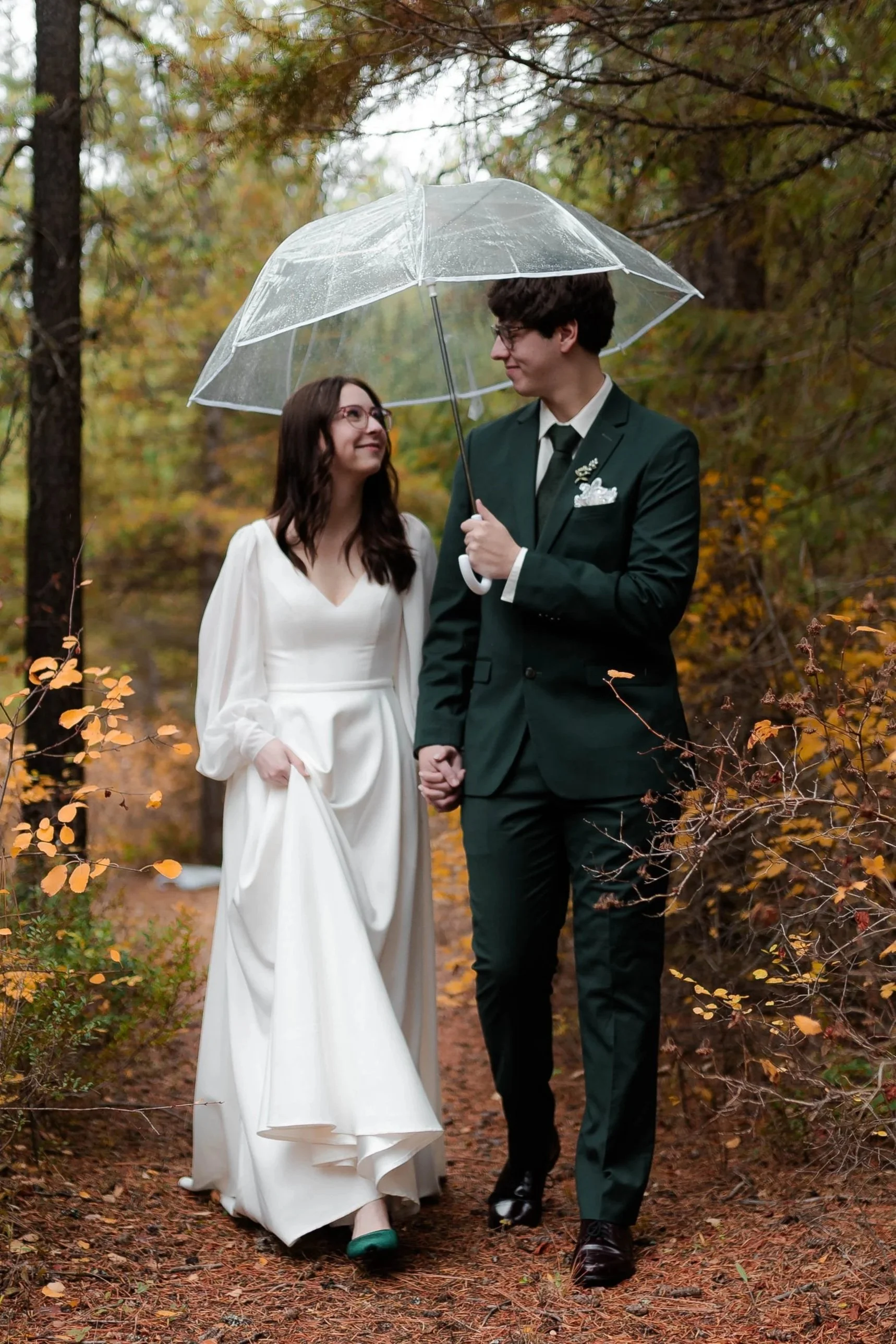 A bride and groom holding hands and standing close under a transparent umbrella on a wooded trail with autumn foliage. An October wedding with rain.