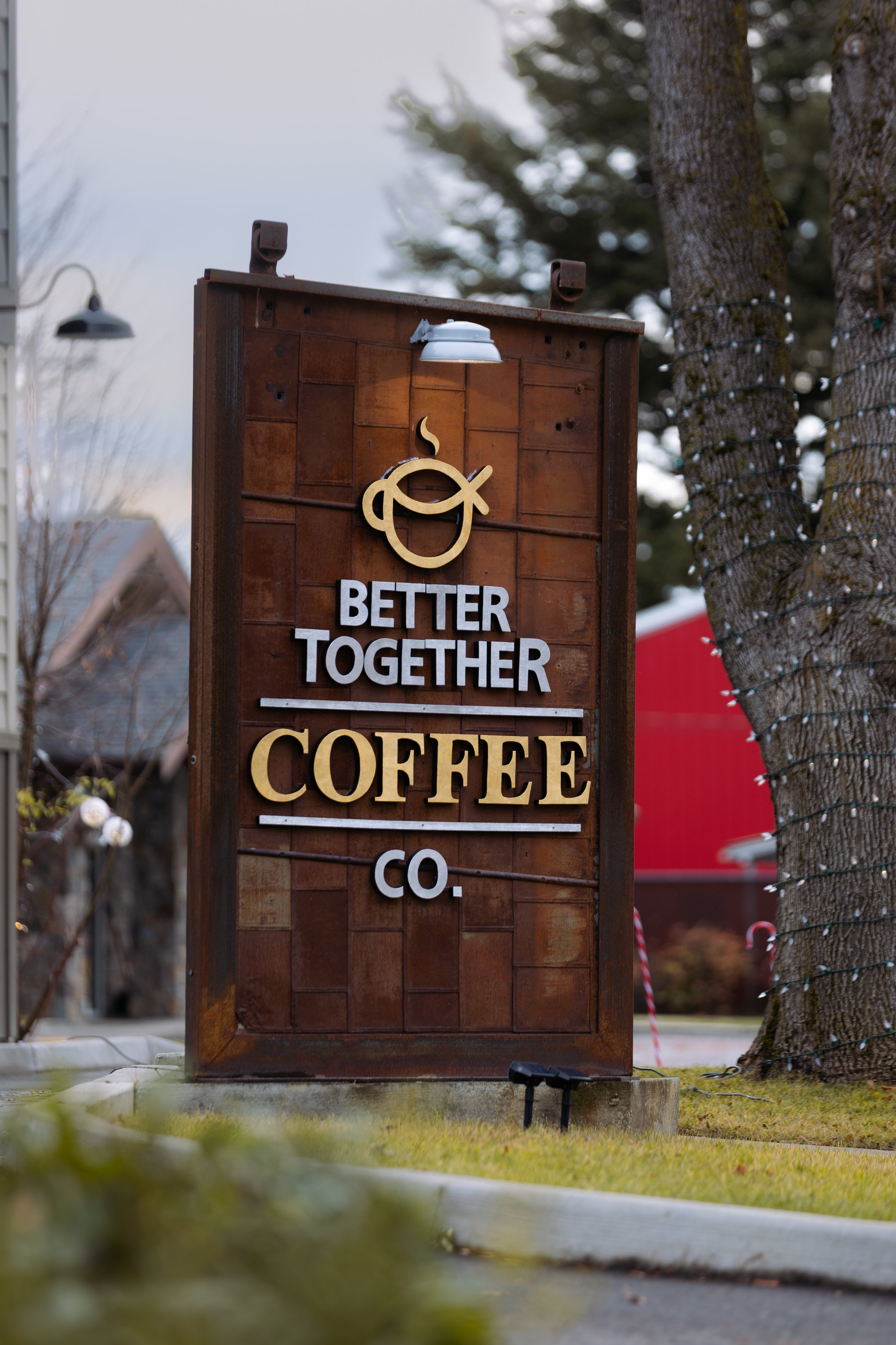 Outdoor signboard with the logo of a coffee cup with steam and the text 'Better Together Coffee Co.' on a wooden background, surrounded by trees and buildings.