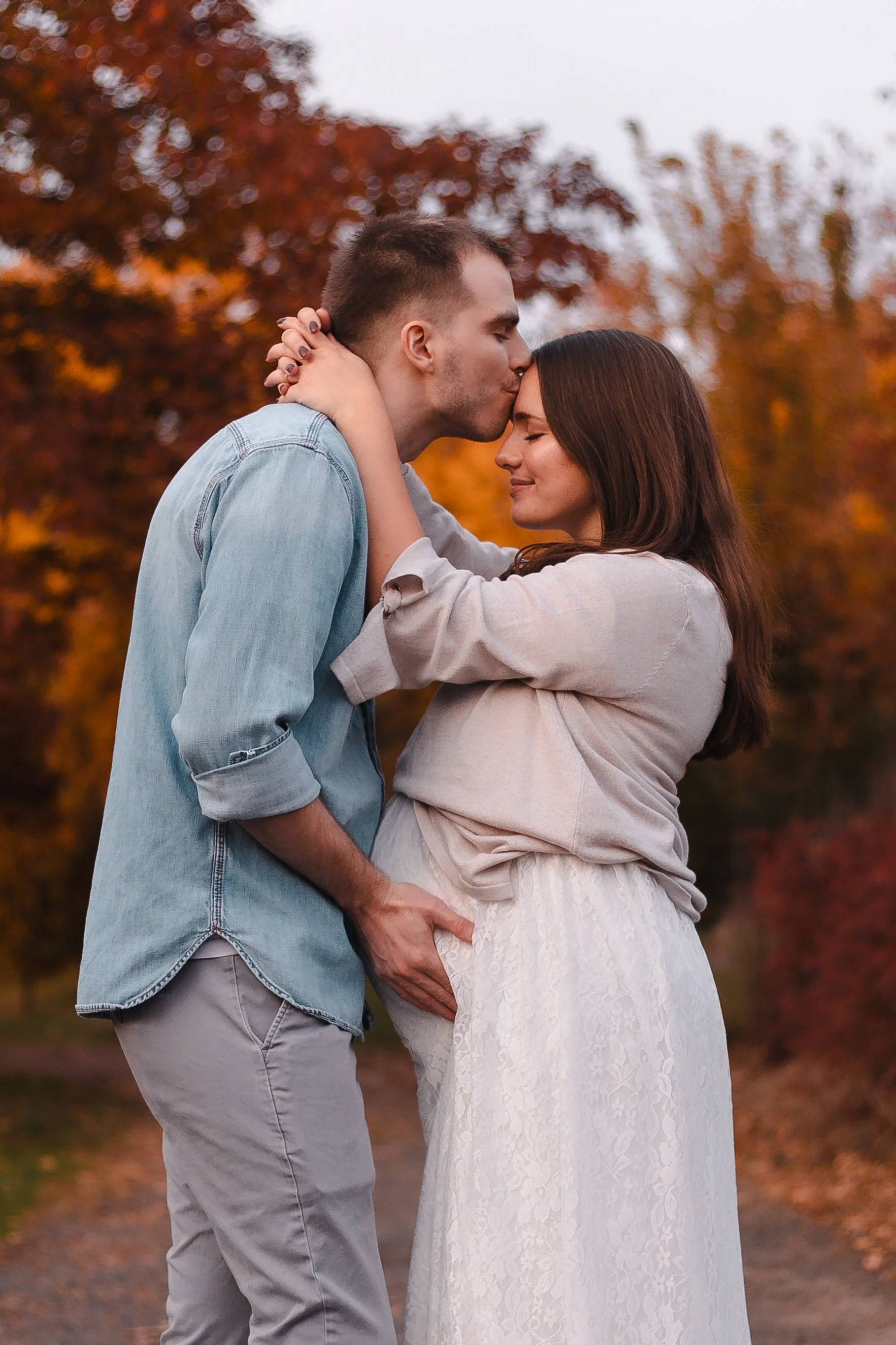 A couple sharing a kiss outdoors during autumn, with the man gently holding the woman's waist and the woman with hands on the man's neck, standing in front of colorful fall foliage.