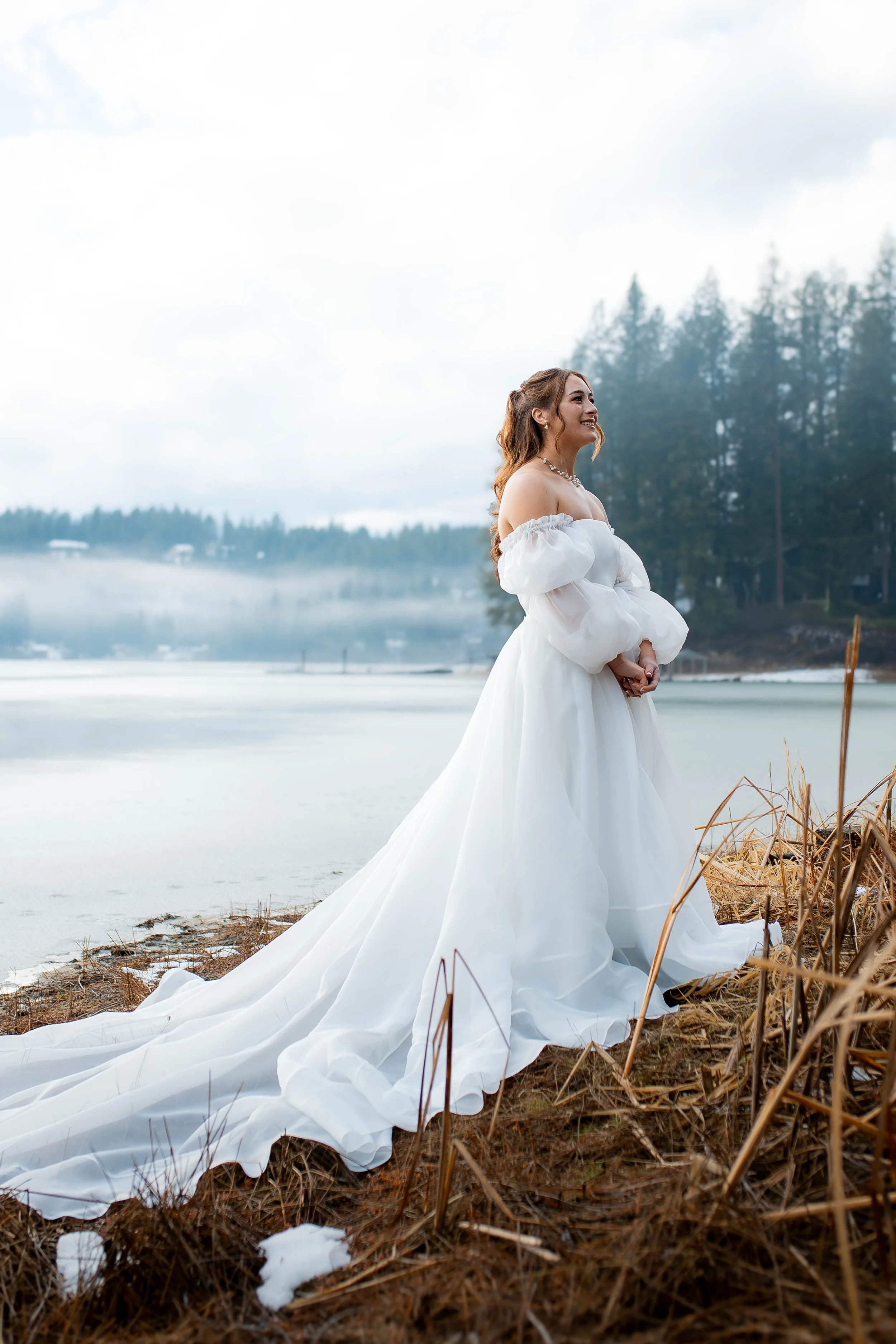 A woman in a white wedding dress standing near a lake with a misty, wooded background.