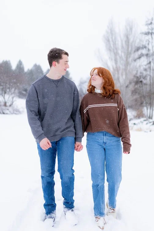 Young couple walking hand in hand through snowy forest on winter day, photographed by LaFleur Photography in Coeur d’Alene.