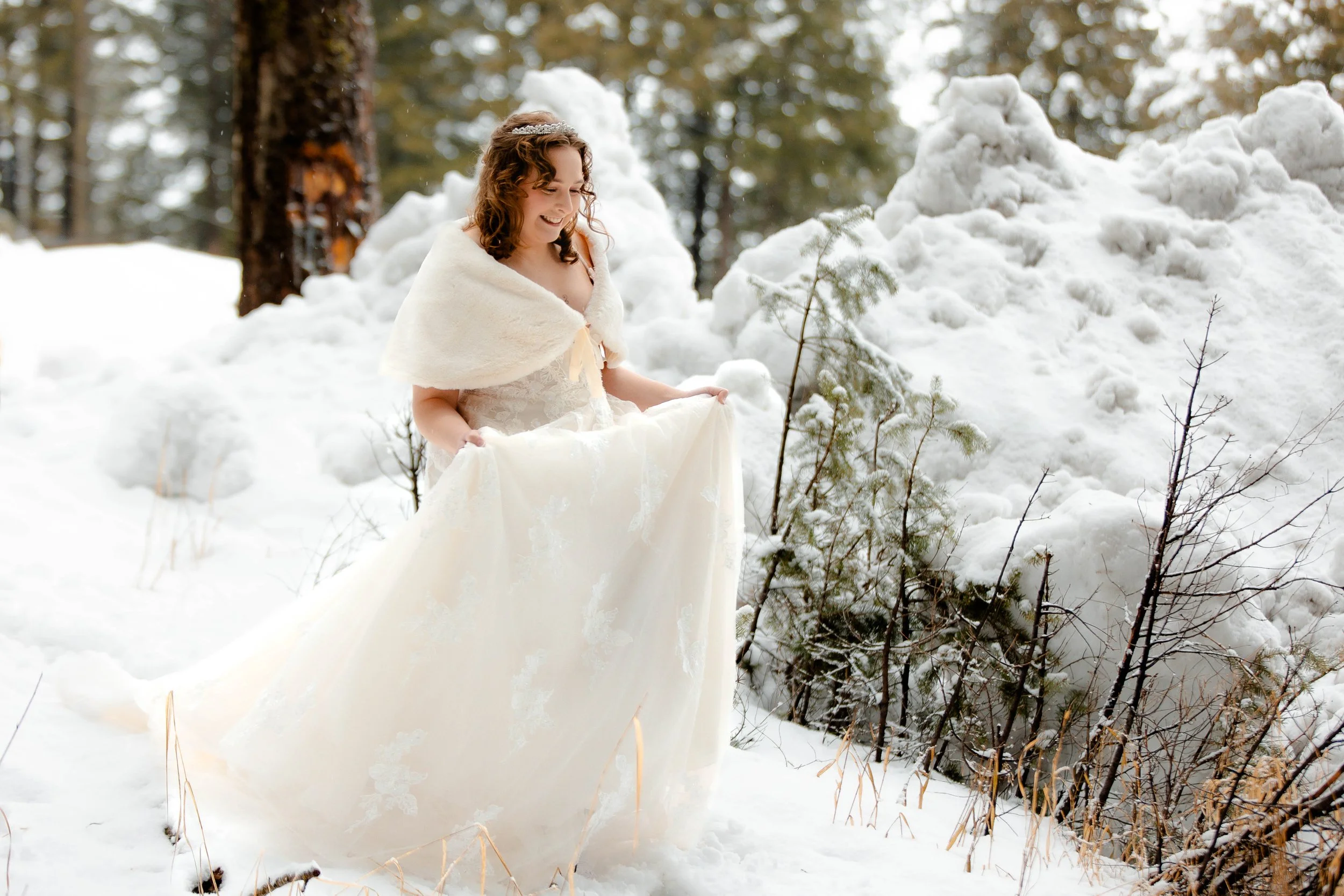 A woman in a wedding dress holding up her gown's skirt in a snowy forest, smiling while looking down. She is about to have her first look with her soon to be husband, at the Scandia Inn, in McCall, ID.