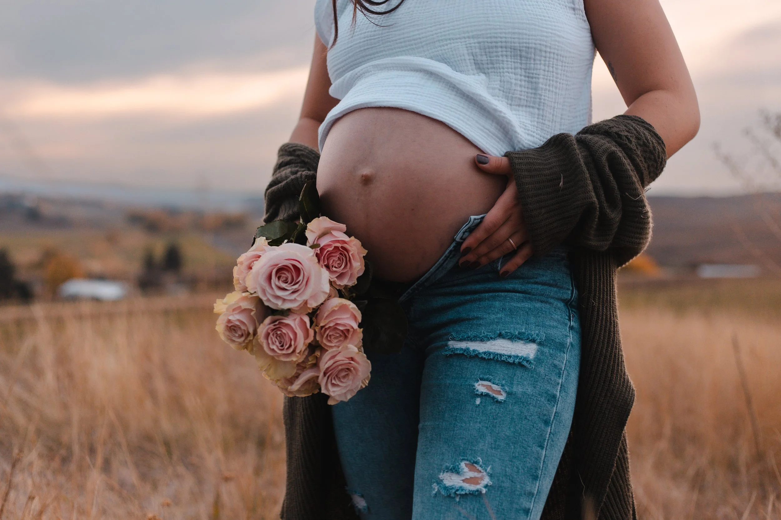 Pregnant woman holding a bouquet of pink roses outdoors during sunset, with field and distant hills in the background.