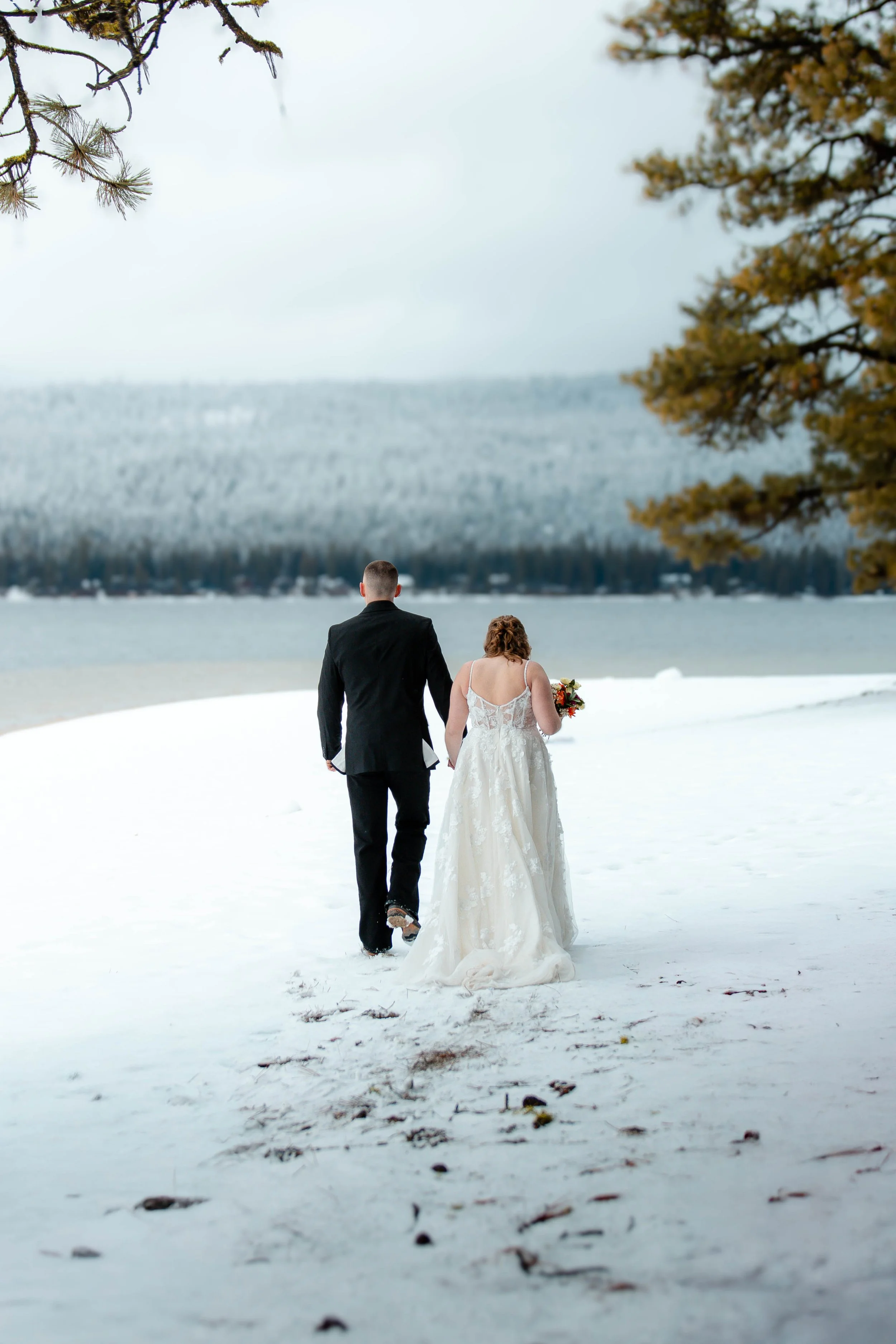 A bride and groom walking hand in hand on a snow-covered lakeshore during winter, with snow-capped mountains and pine trees in the background, at Ponderosa State Park in McCall, ID after just being eloped.