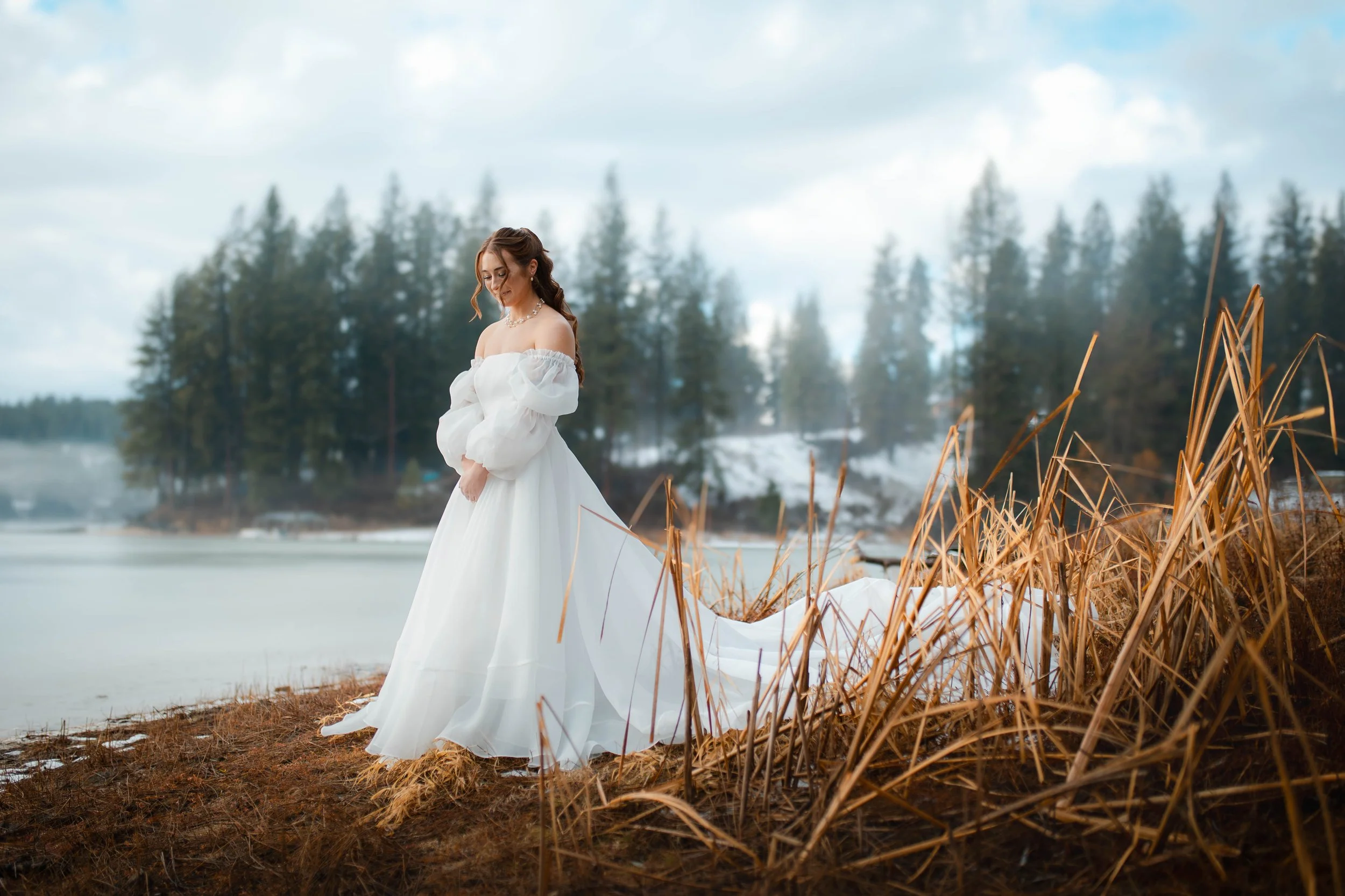 A woman in a white off-the-shoulder wedding dress with puffy sleeves standing on a forested lakeshore, with tall dry grasses and a lake in the background. At the Hayden Lake Lodge, in Hayden, ID. 