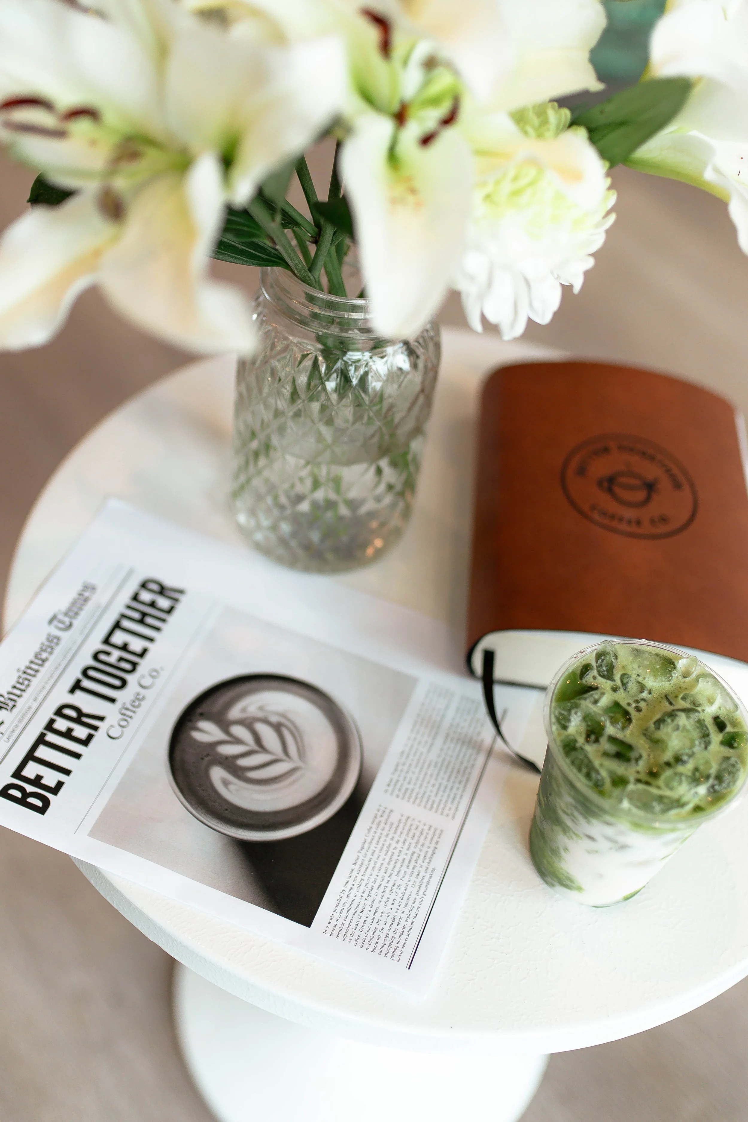 A white round table with a vase of white flowers, a newspaper, a brown leather-bound notebook, and a glass of green and white beverage with ice and mint leaves for Better Together Coffee Co in Coeur d'Alene, ID.
