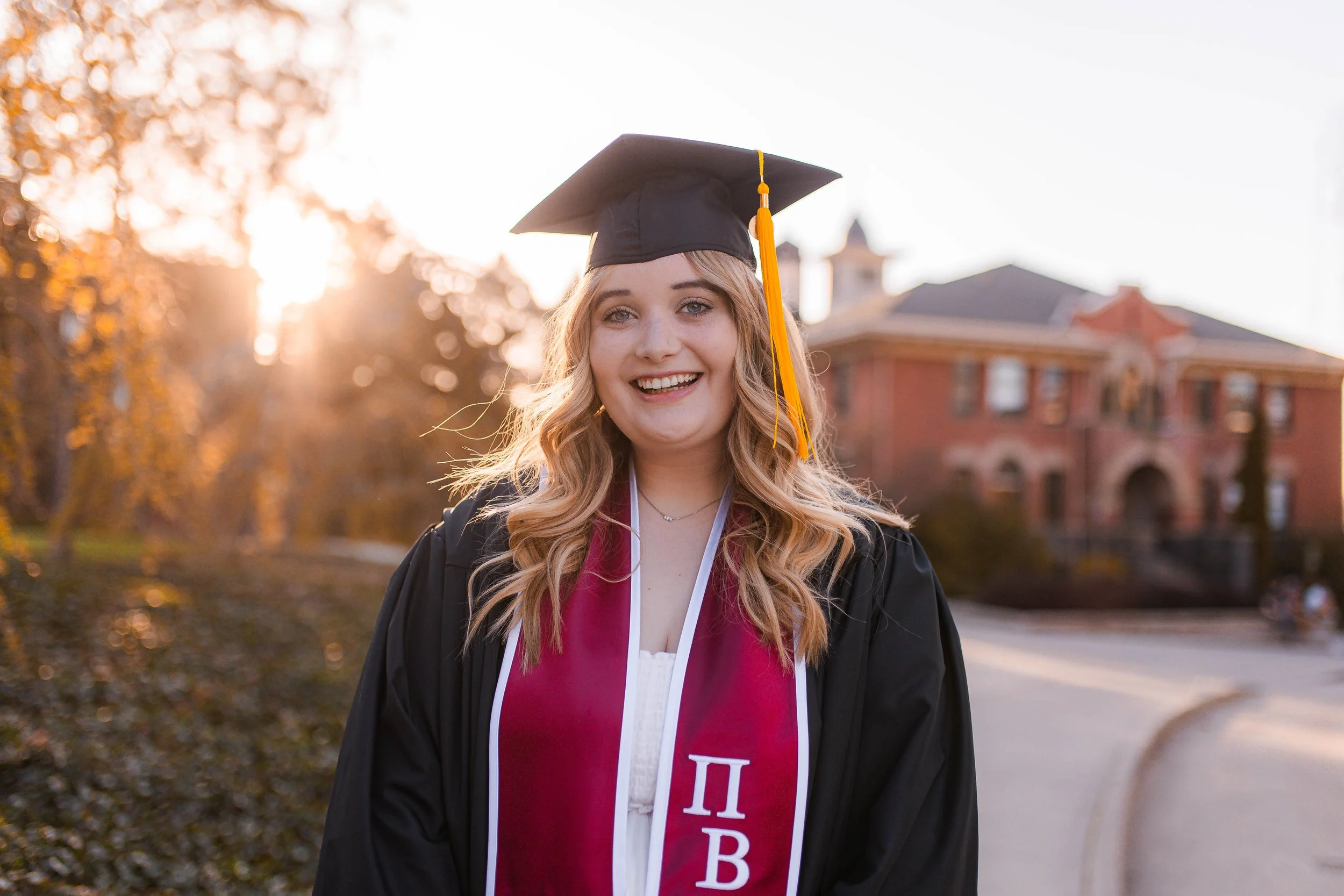 Senior portraits & Cap and gown portraits. Don't miss out on grad shoots! A smiling young woman in graduation cap and gown standing outdoors with a university building in the background during sunset.