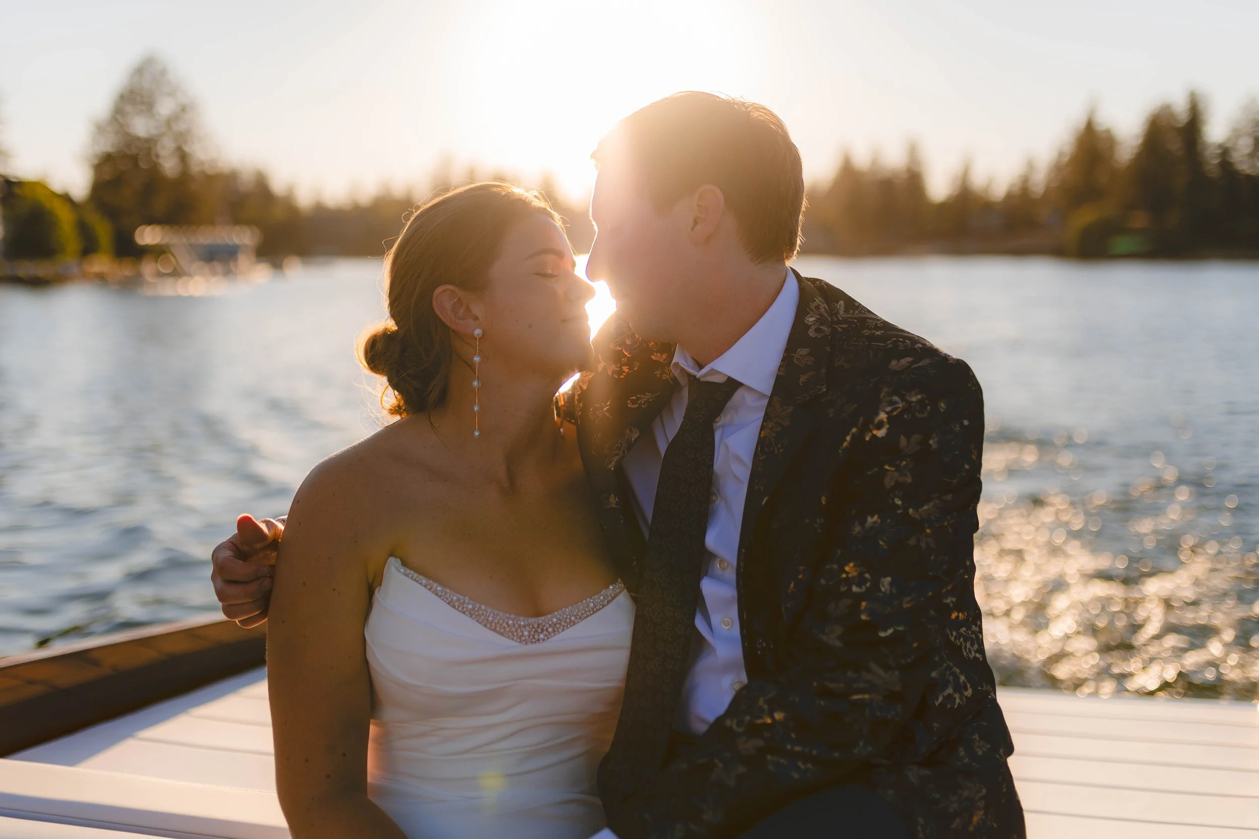 A couple dressed in wedding attire, sitting close together on a boat during sunset, engaging in an intimate moment by a body of water with trees in the background. Wedding photo on Lake Coeur d' Alene.