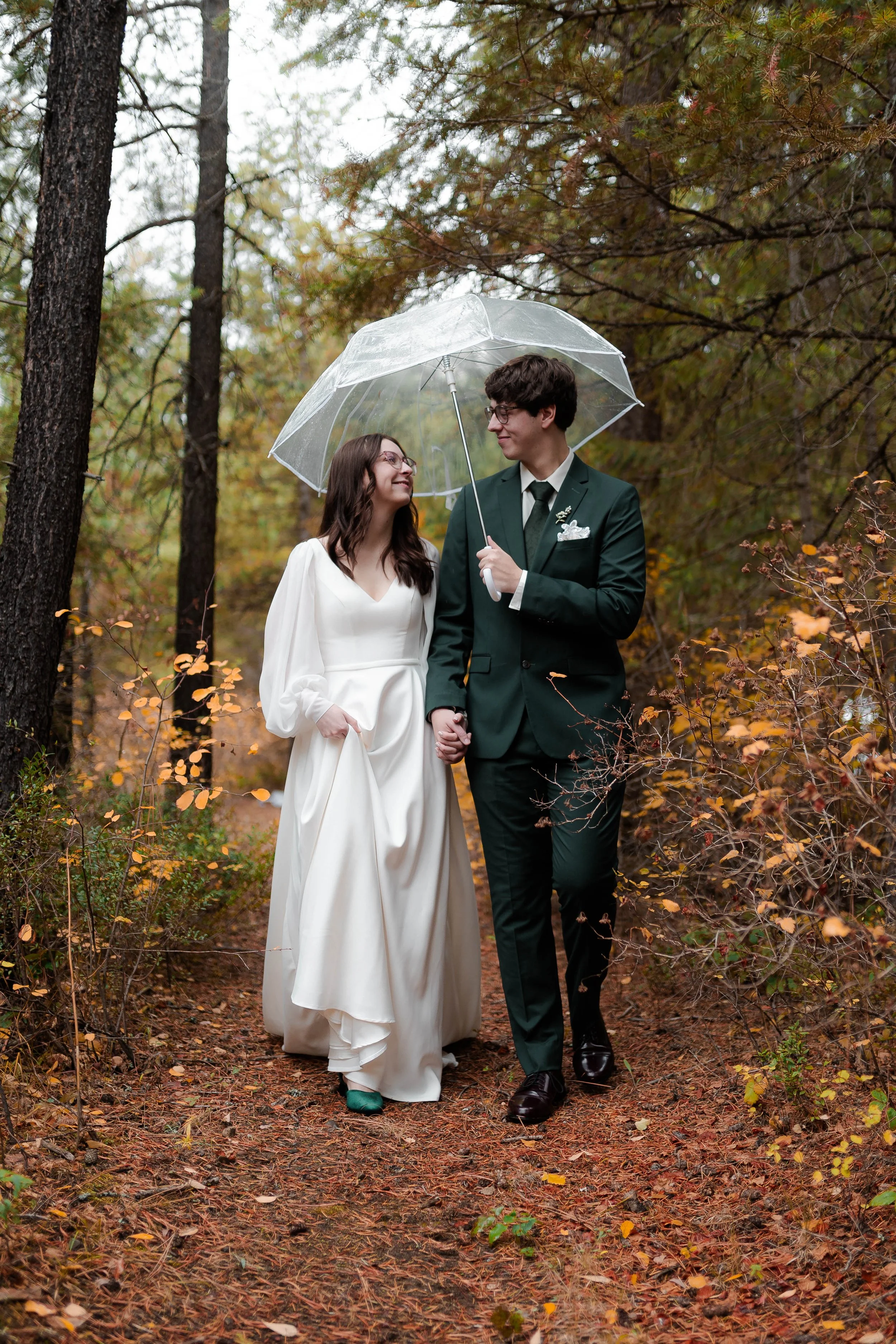 A bride and groom holding hands under a transparent umbrella walk through a wooded area with autumn leaves.