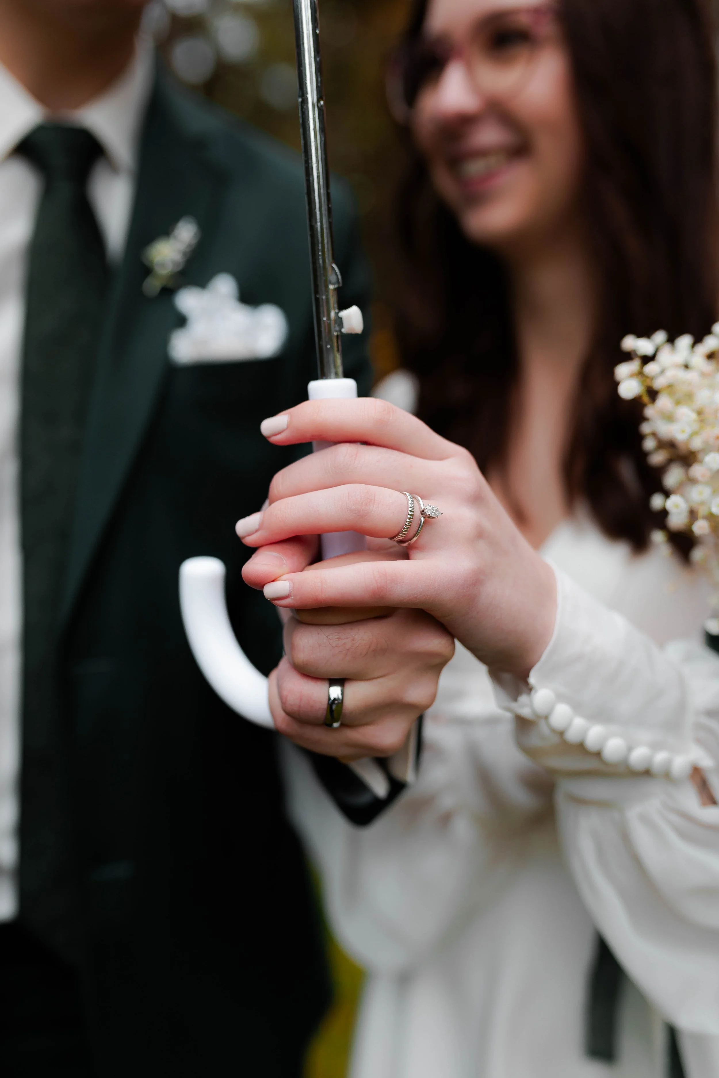 Close-up of a woman holding a white umbrella, wearing a ring with a diamond on her finger. She is smiling and holding a bouquet of small white flowers. A man in a dark suit and tie stands behind her, partially visible.