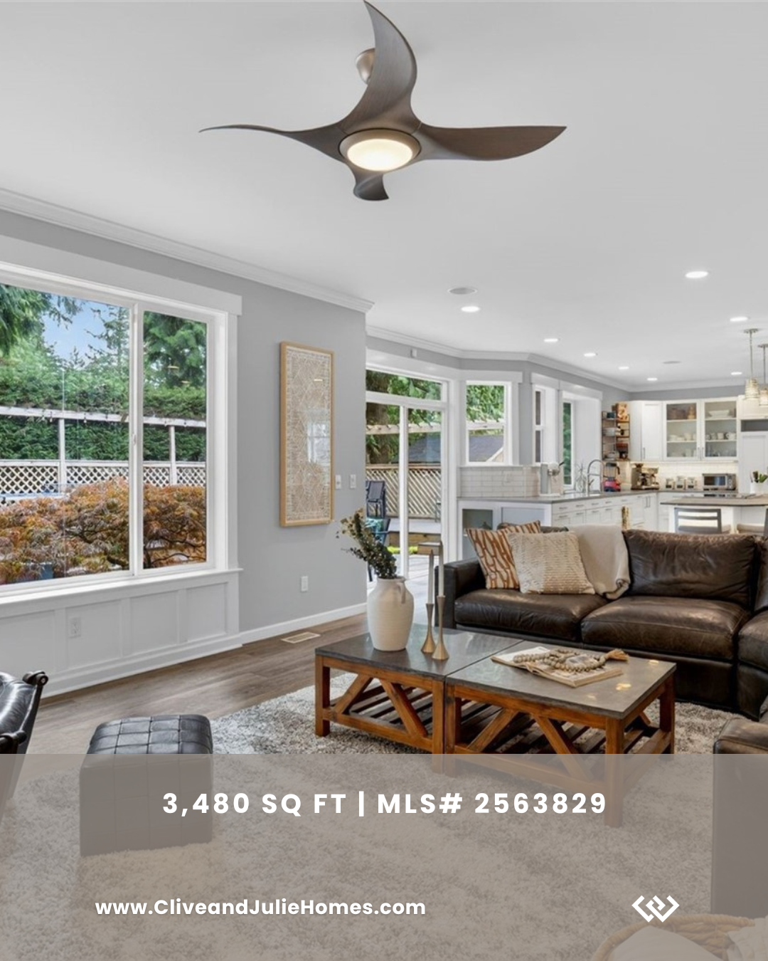 Living room with large windows, gray walls, modern ceiling fan, leather sofa, wooden coffee table, decorative vases, and a view of the backyard.