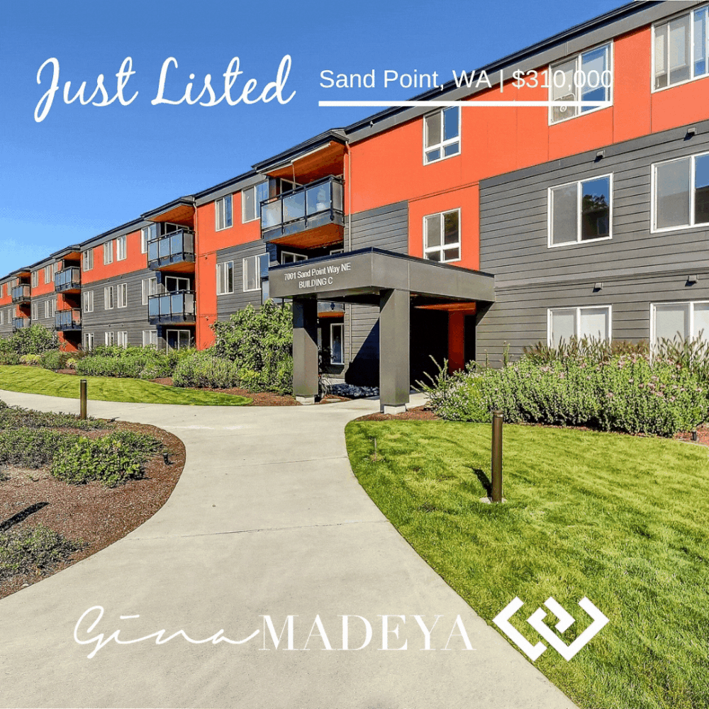 A multi-story apartment building with gray and red exterior walls, balconies, and an entrance canopy, surrounded by a well-maintained lawn and garden, under a clear blue sky.