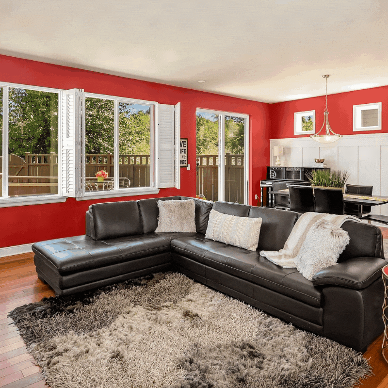 Living room with red walls, large windows with white shutters, black leather sectional sofa, shaggy rug, and a dining area with a black table and chairs in the background.
