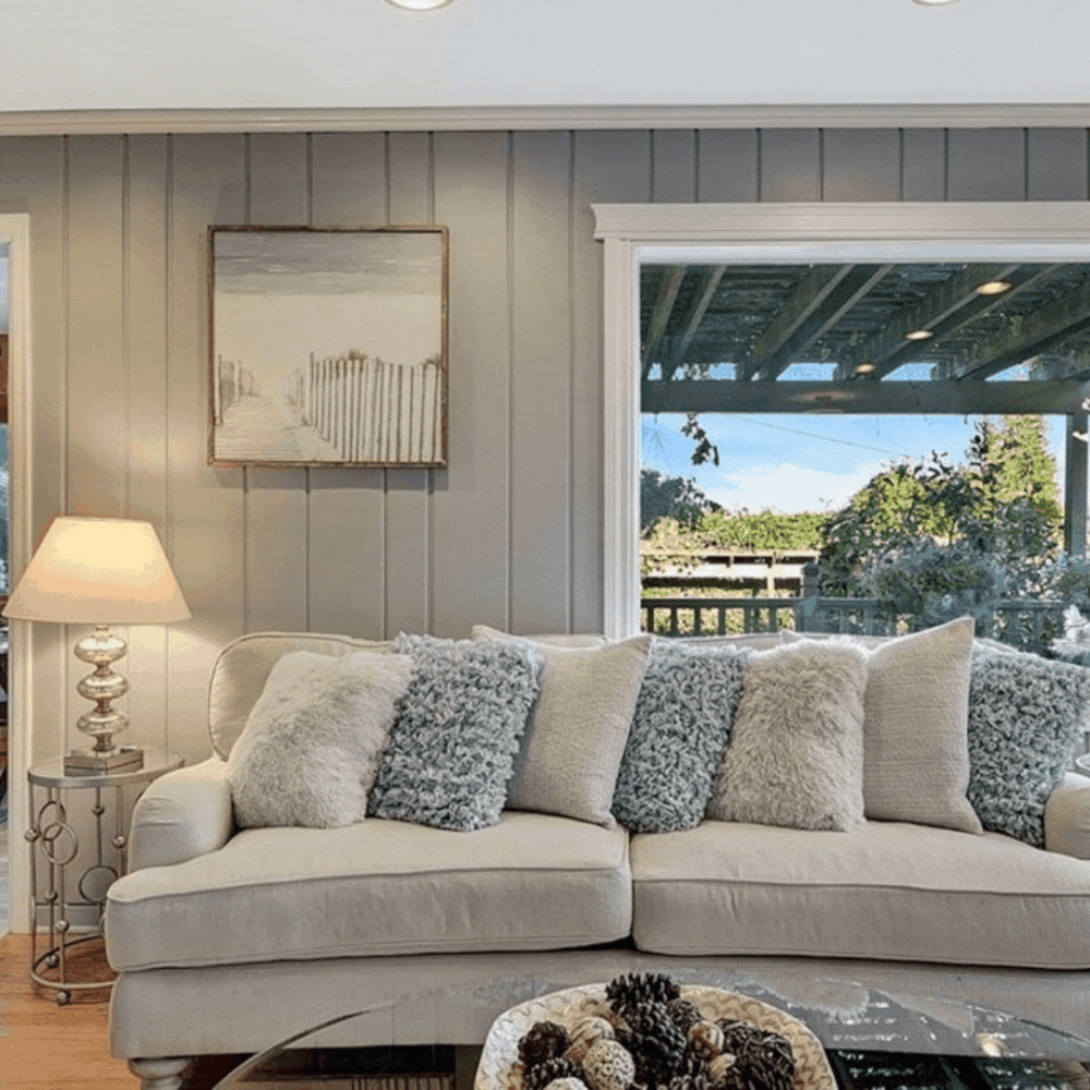 Living room with beige sofa, multiple textured throw pillows, a side table with a lamp, and a large window showing a backyard with trees and a deck.
