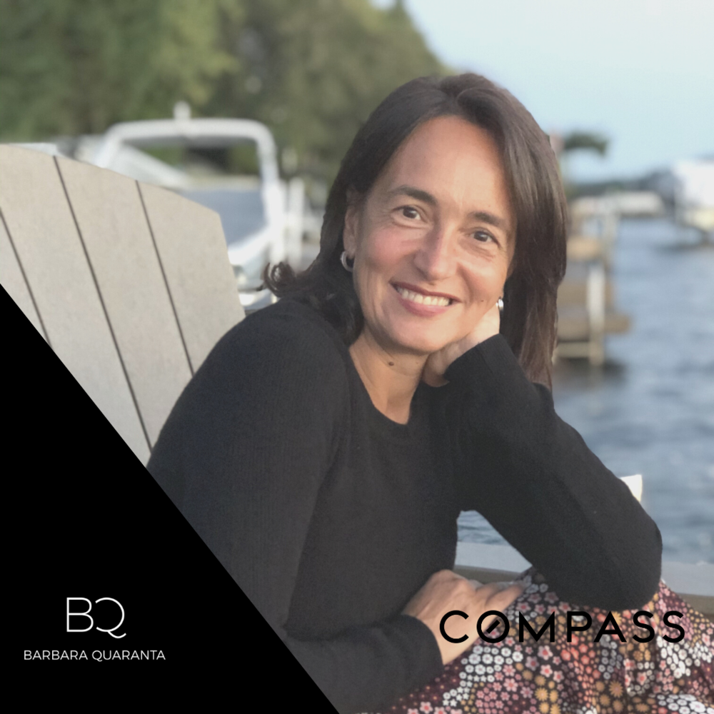 A woman with shoulder-length brown hair smiling, sitting outdoors near a body of water during the day, with boats and trees in the background.