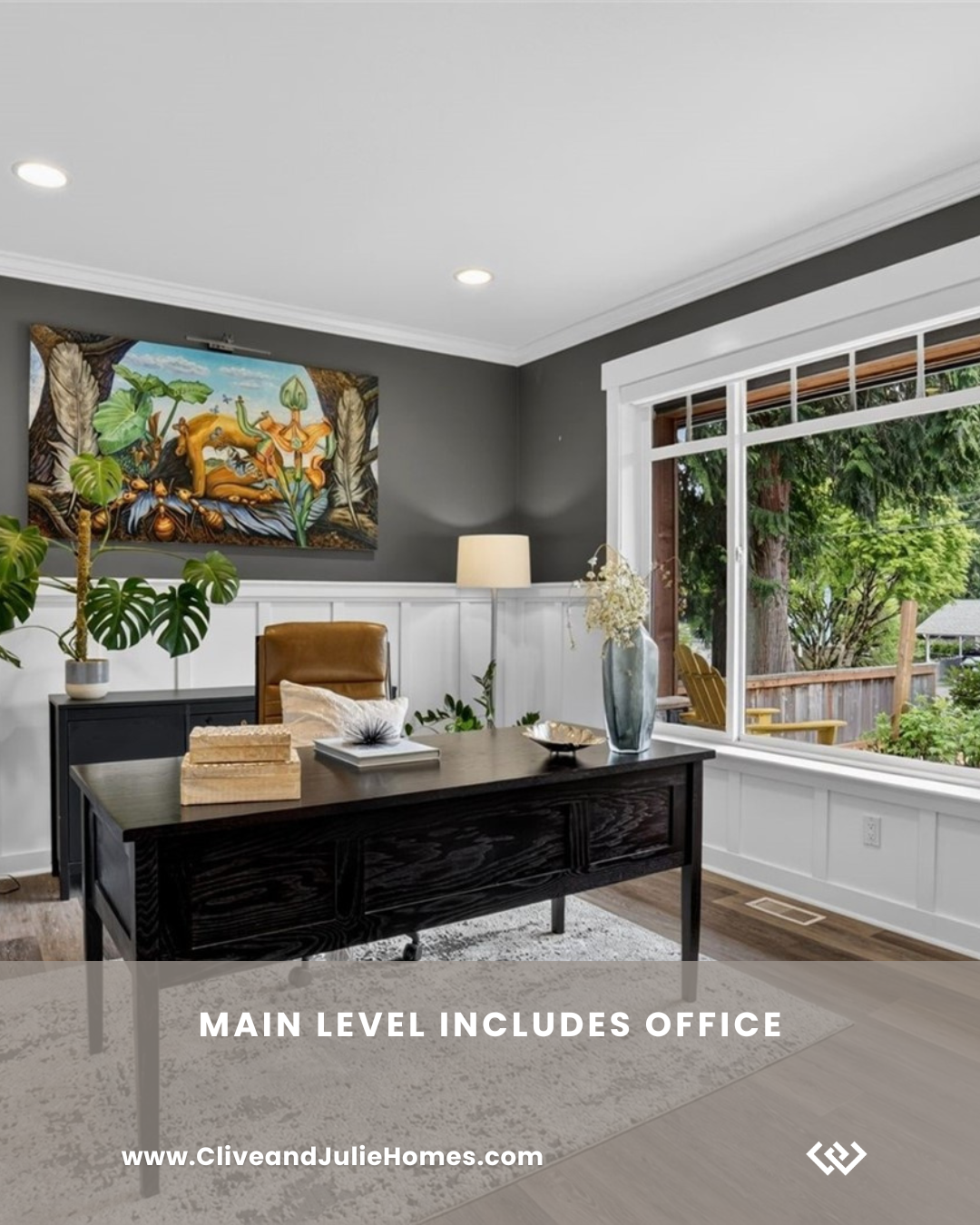 Main level home office with a dark wooden desk, tan leather chair, large window showing outdoor greenery, a lamp, a large vase with white flowers, a painting on the wall, and a white and beige area rug.