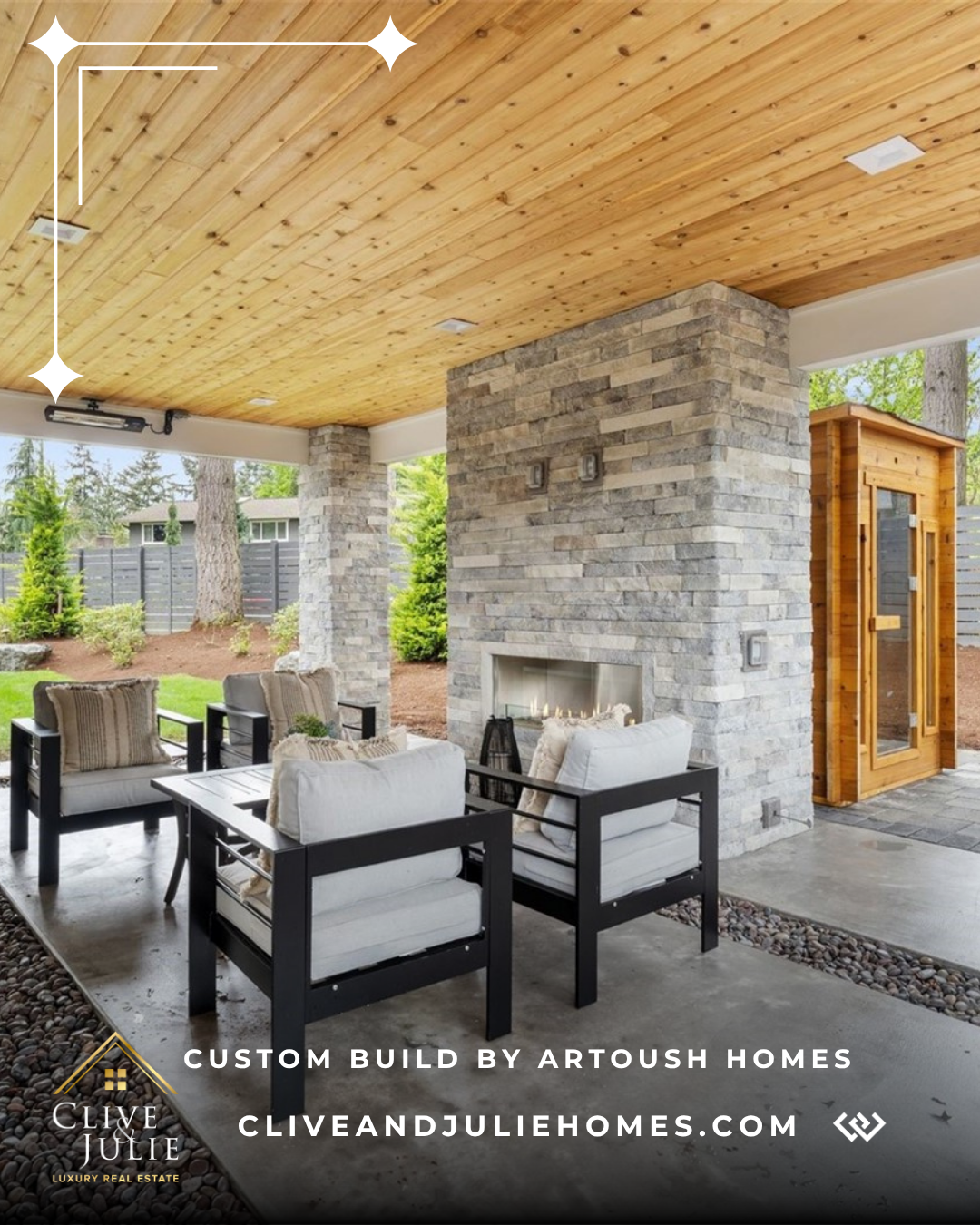 Covered outdoor patio with a wood-paneled ceiling, stone fireplace, black outdoor furniture with white cushions, and a wooden sauna in the background. Green trees and a gray fence are visible beyond.