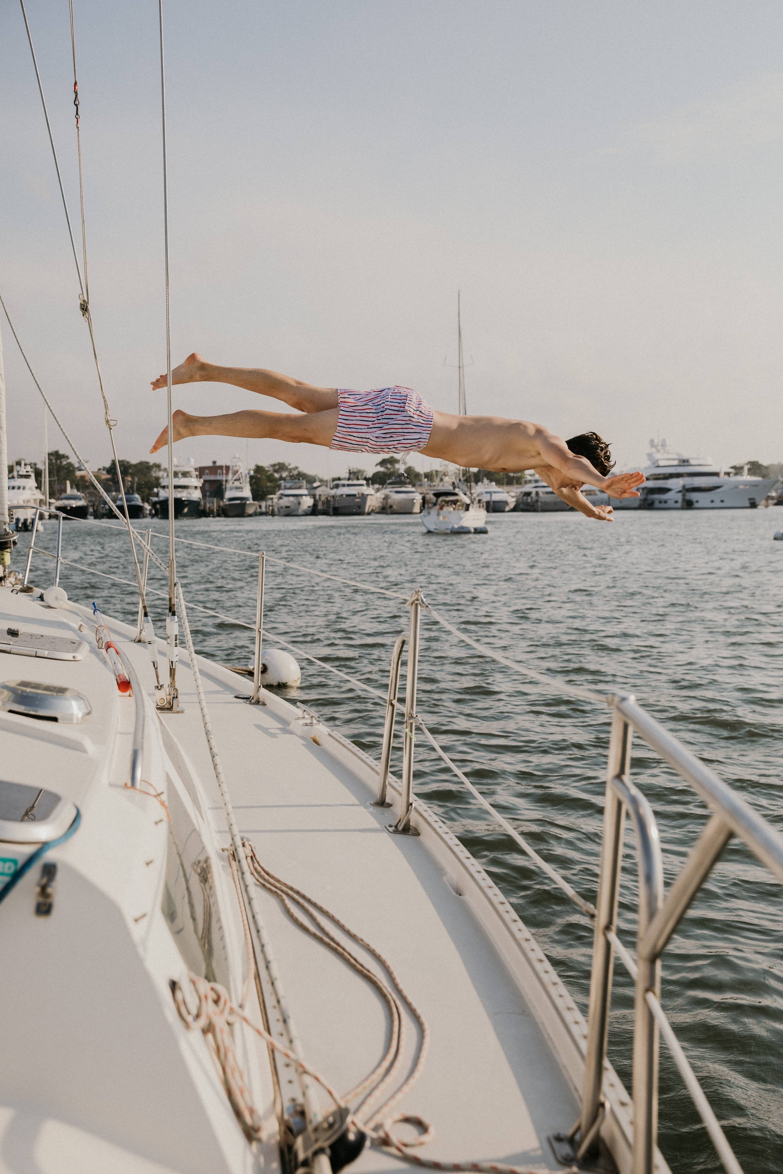 A person in striped shorts is diving off the side of a boat into the water near a marina with boats docked, under a partly cloudy sky.