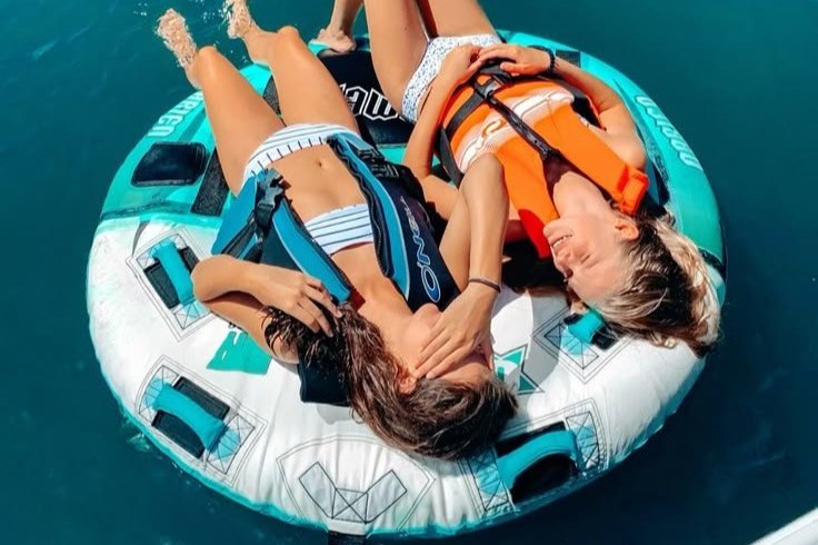 Two young women in swimsuits and life jackets lying on a blue and white inflatable raft floating in the water.