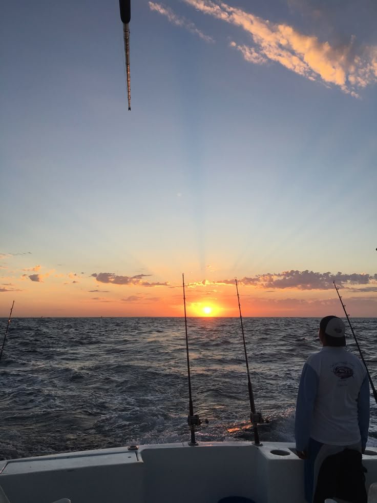 A person on a boat fishing during sunset, with three fishing rods set up and an ocean view in the background.