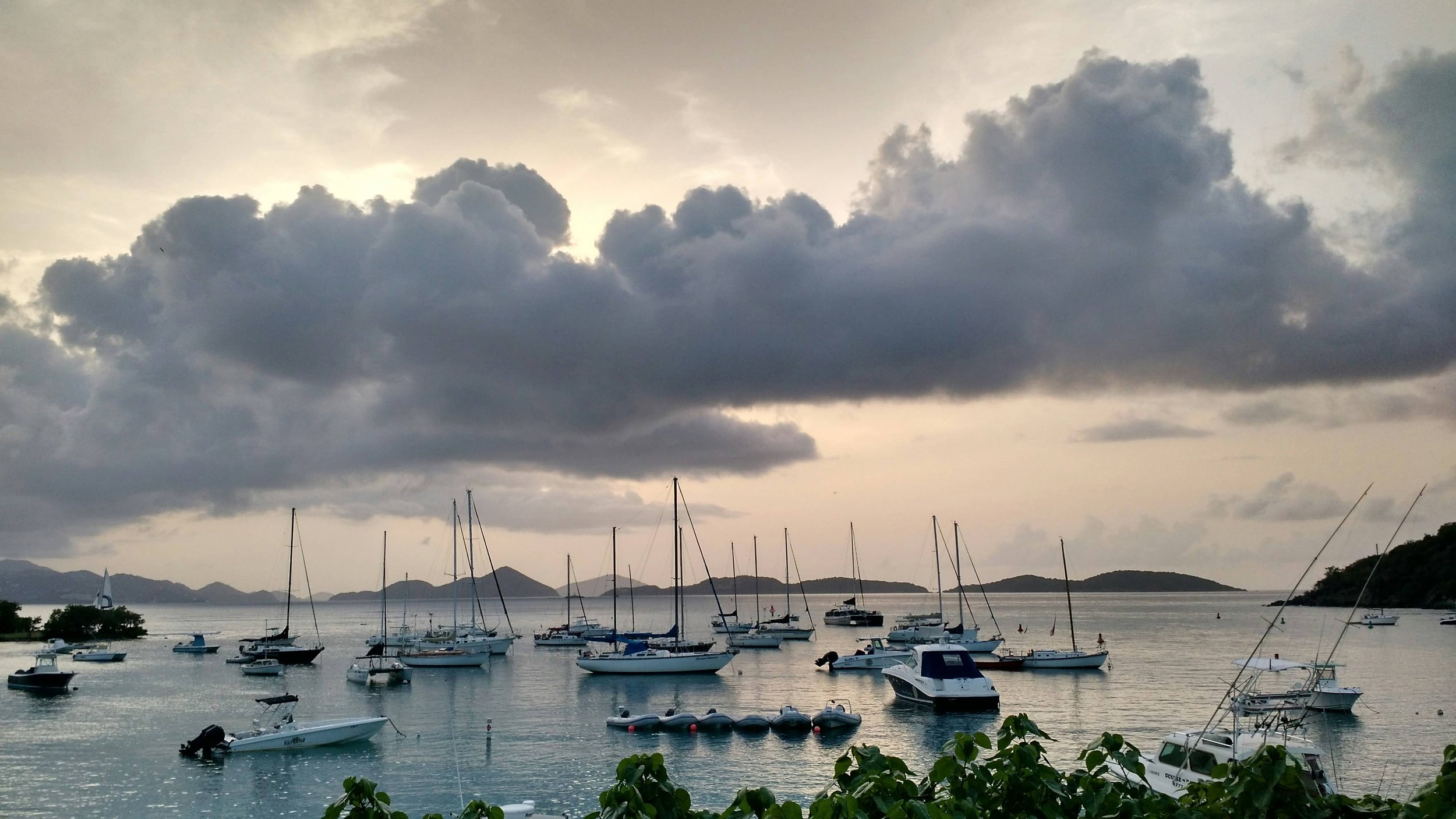 A marina with numerous sailboats and motorboats in calm water, hills in the distance, and a cloudy sky overhead.