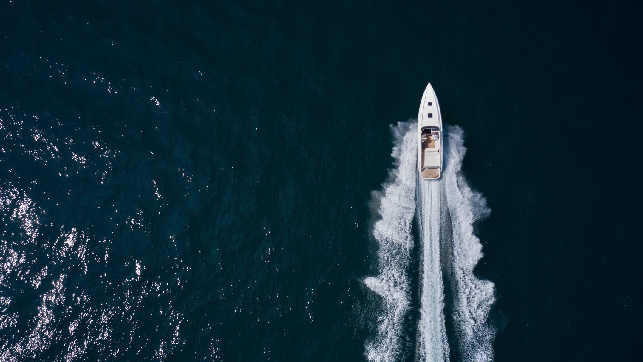 Aerial view of a white speedboat moving through the dark blue ocean, creating white wake behind it.
