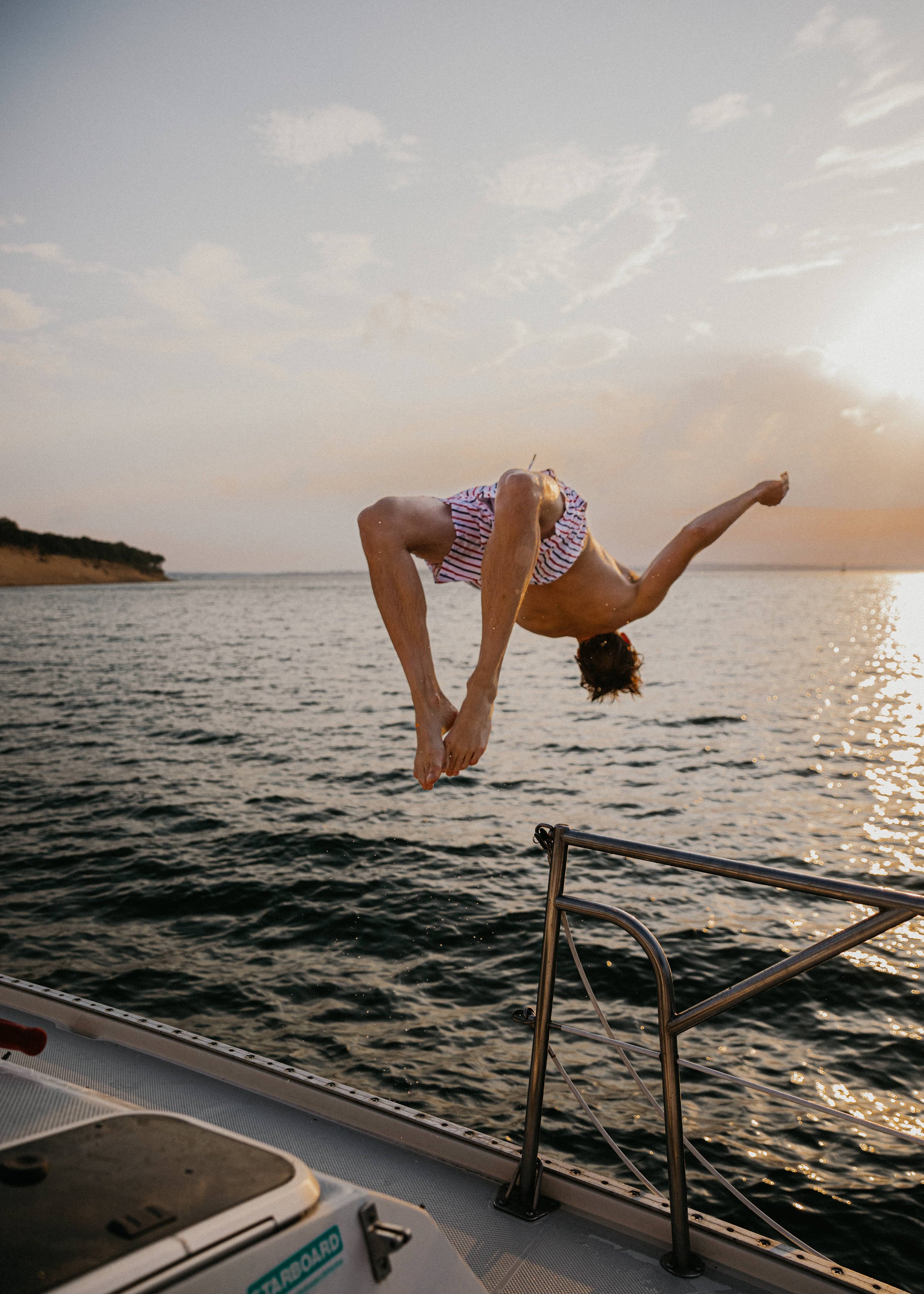 A person performing a backflip or somersault off the edge of a Sailboat into a Sag Harbor during sunset, with the water and sky in the background.