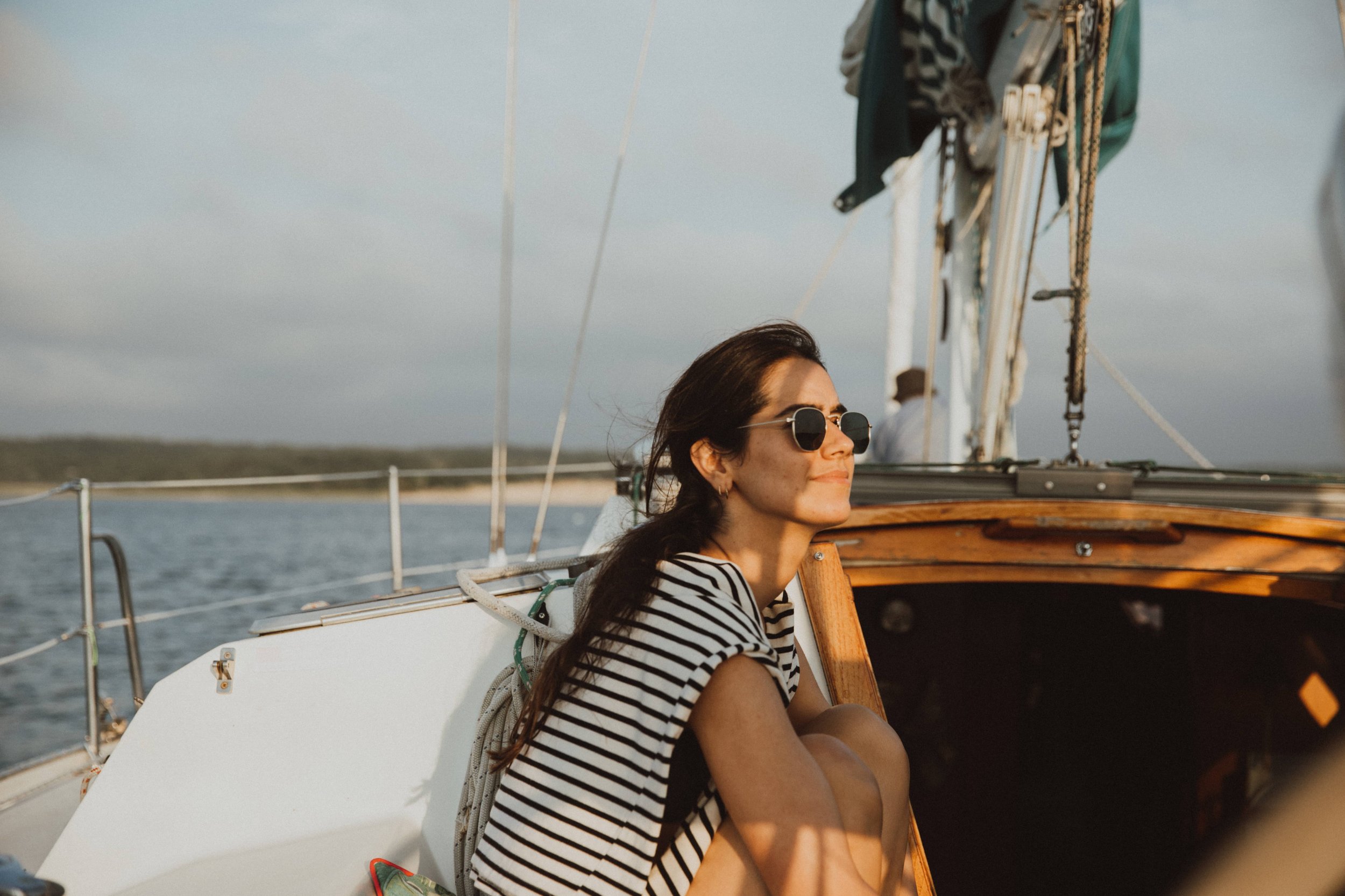 A woman wearing sunglasses and a black-and-white striped shirt sitting on a boat with a smiling expression, with water, a shoreline, and a cloudy sky in the background.