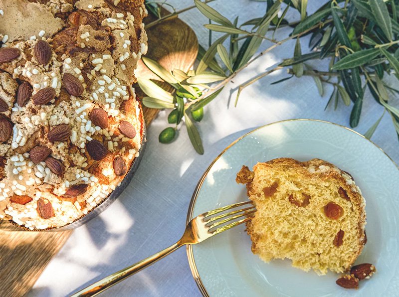 Een stuk cake met rozijnen op een bord en een vork ernaast, naast een grote cake met noten en suikerglazuur, op een witte tafel met groen blad