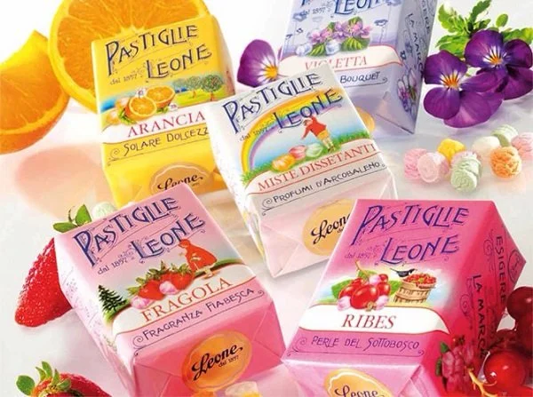 Assortiment van kleurrijke pastilles en snoepjes, verpakt in vrolijke doosjes met afbeeldingen van fruit en bloemen, naast verse bloemen en fruit.