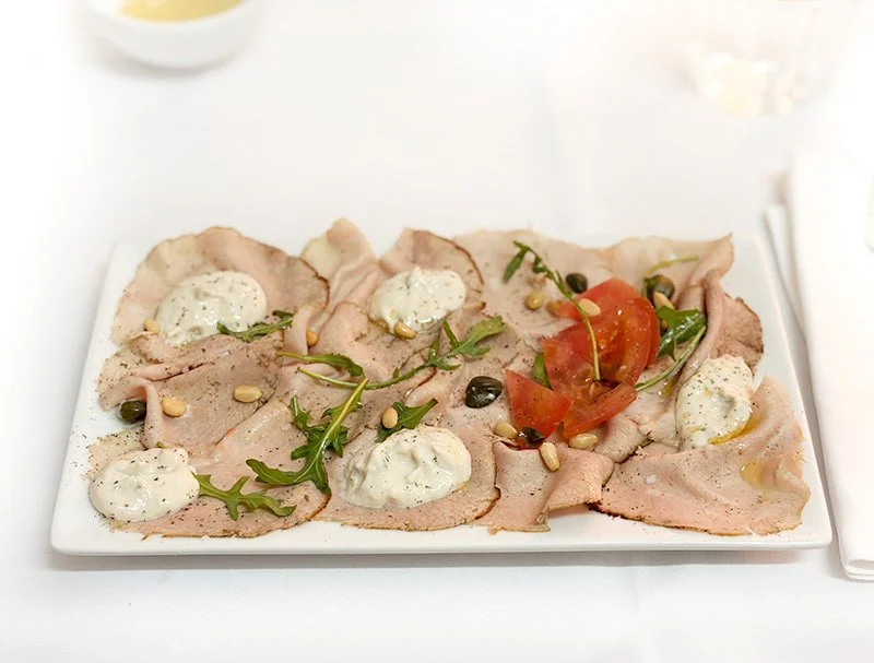 Een bord met dun gesneden vlees, gegarneerd met saus, tomaat, rucola, en pine nuts.
