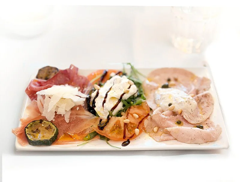 Mediterraan antipasto schaal met geitenkaas, ham, courgette, tomaat, kaas, balsamico siroop, pijnboompitten en kappertjes op een witte rechthoekige schaal.